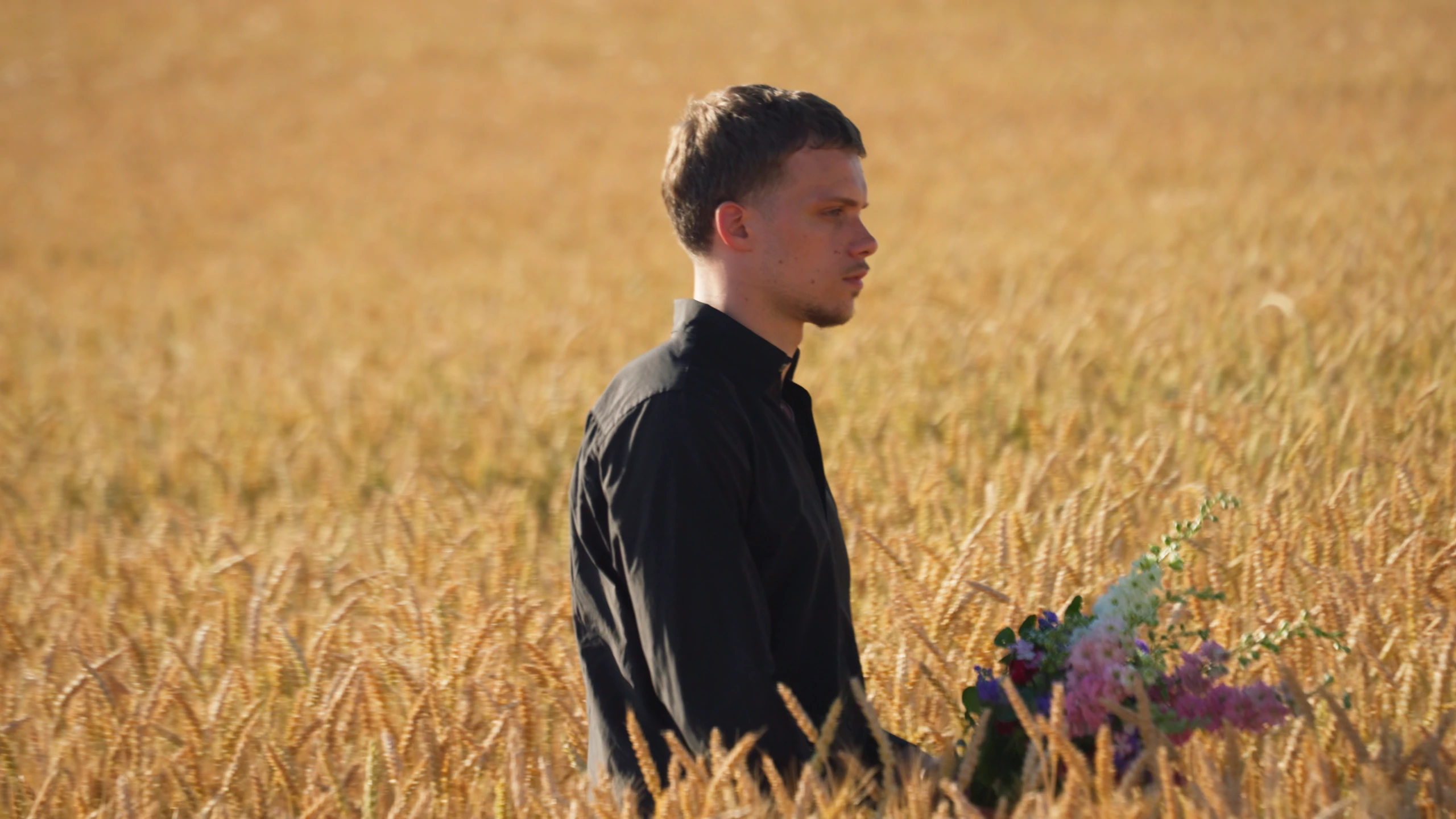 Un homme en chemise noire debout dans un champ de blé, un bouquet de fleurs à la main.