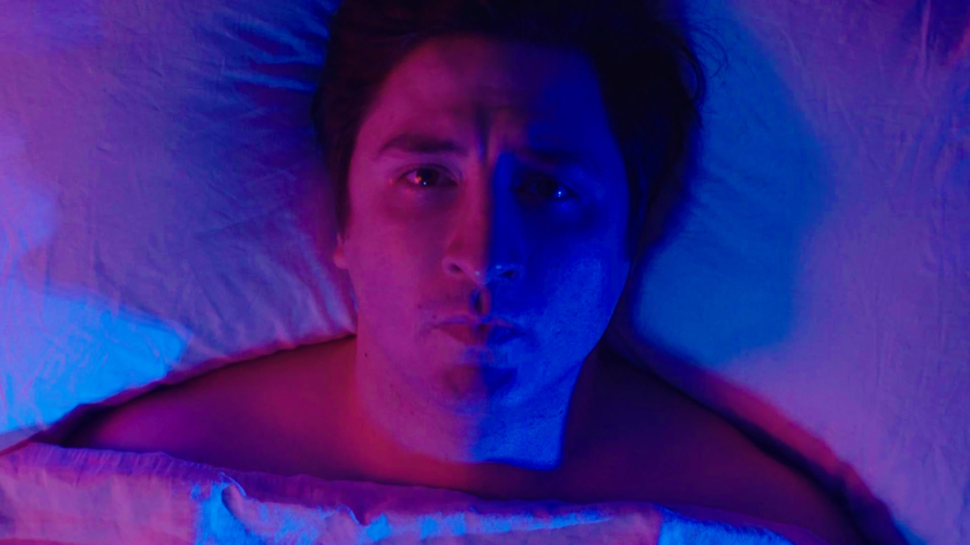 Un homme allongé dans son lit, le visage partagé entre une lumière rouge et une lumière bleue.