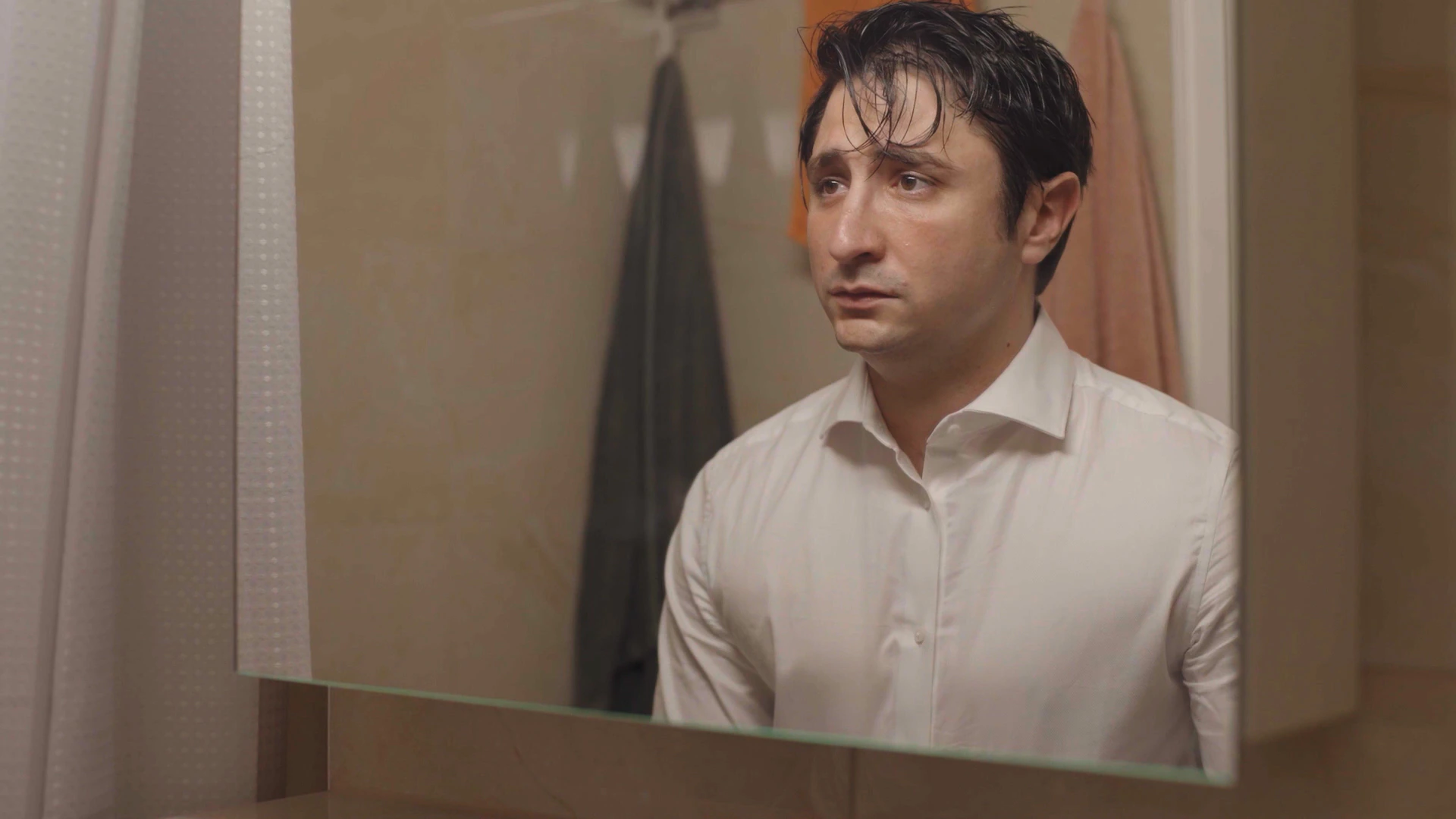 Un homme aux cheveux mouillés et en chemise blanche se regarde, inquiet, dans le miroir de sa salle de bains.