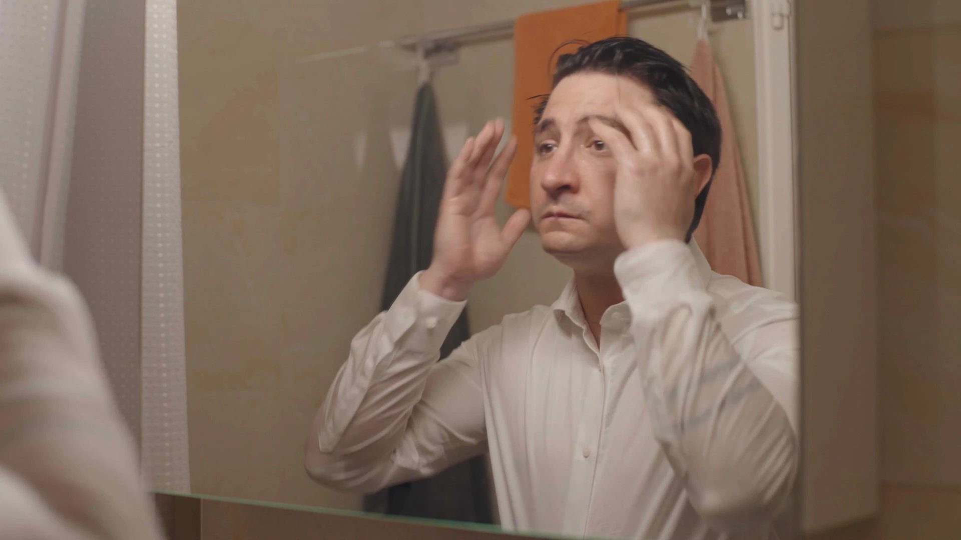 Un homme en chemise blanche se passe les mains dans les cheveux devant le miroir de la salle de bains.