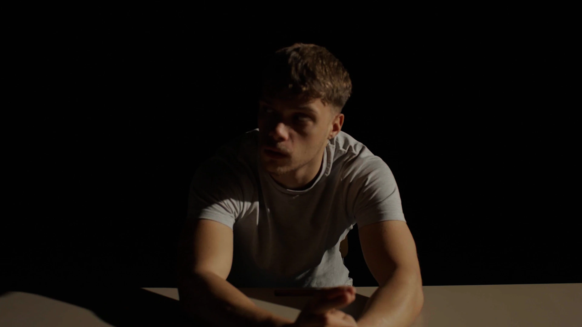 Un jeune homme en t-shirt gris assis à une table, penché en avant, seul dans le noir.