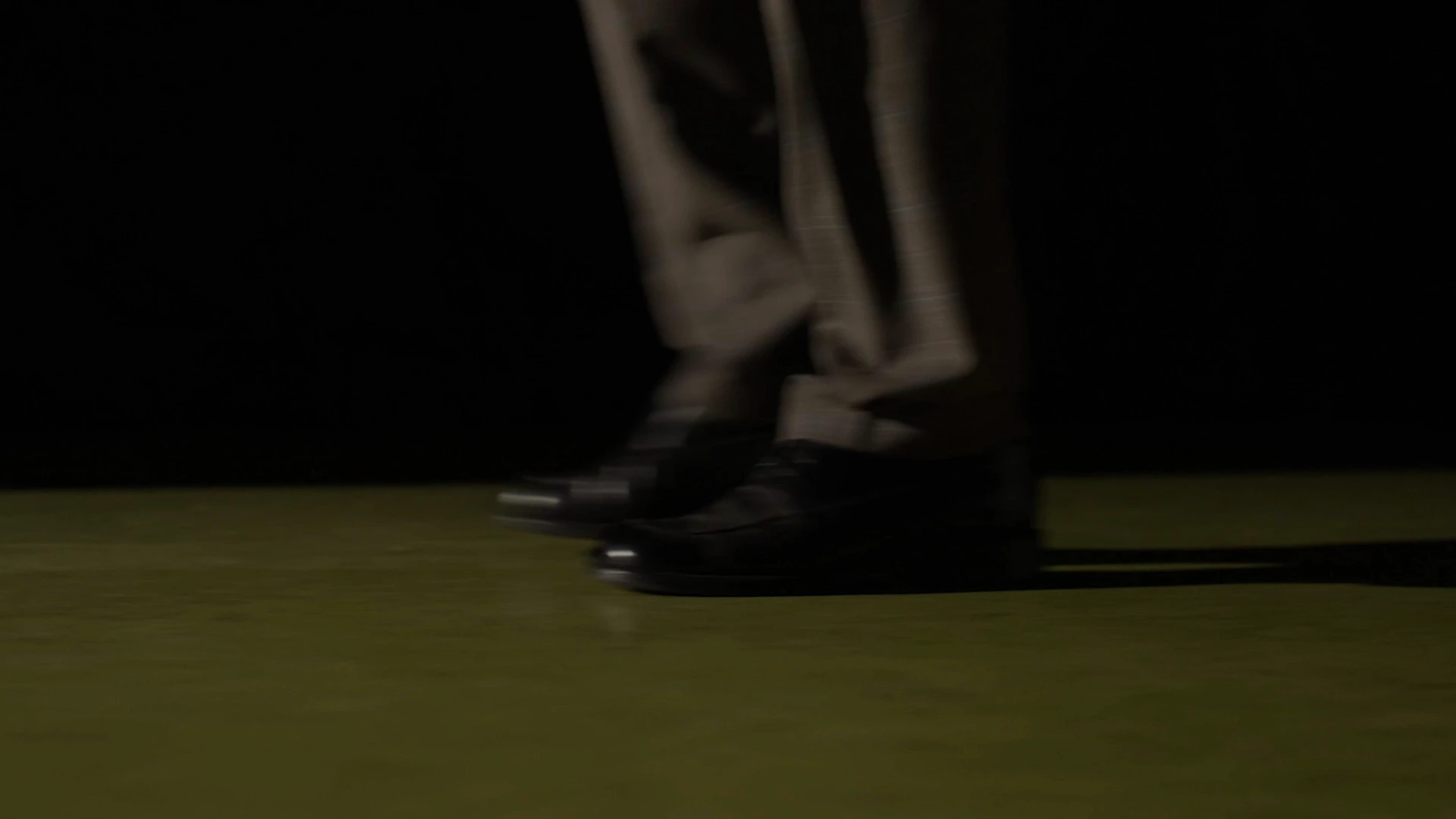 Les jambes d'un homme en costume et ses chaussures noires sur un sol vert, entourées de noir.