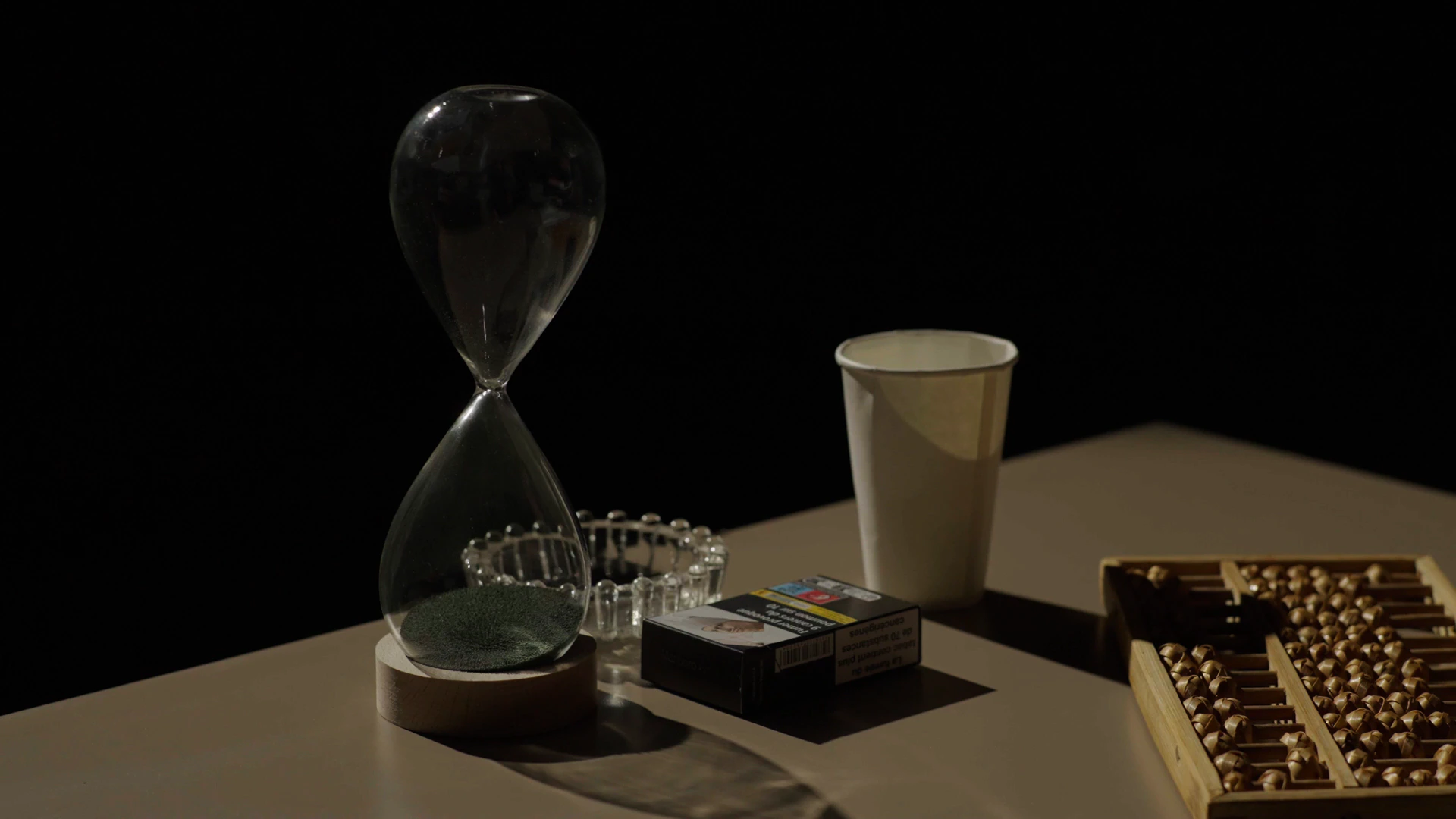 Sur une table dans l'obscurité, un sablier, un cendrier, un gobelet en carton et un boulier.