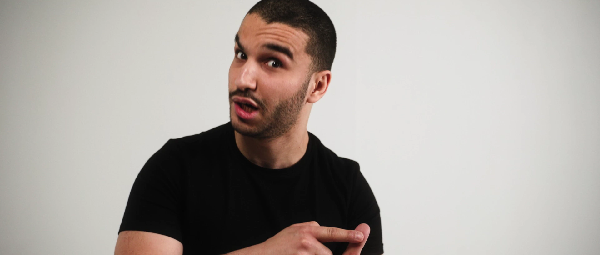 Un homme en t-shirt noir, sur fond blanc, parle à la caméra en comptant sur ses doigts.