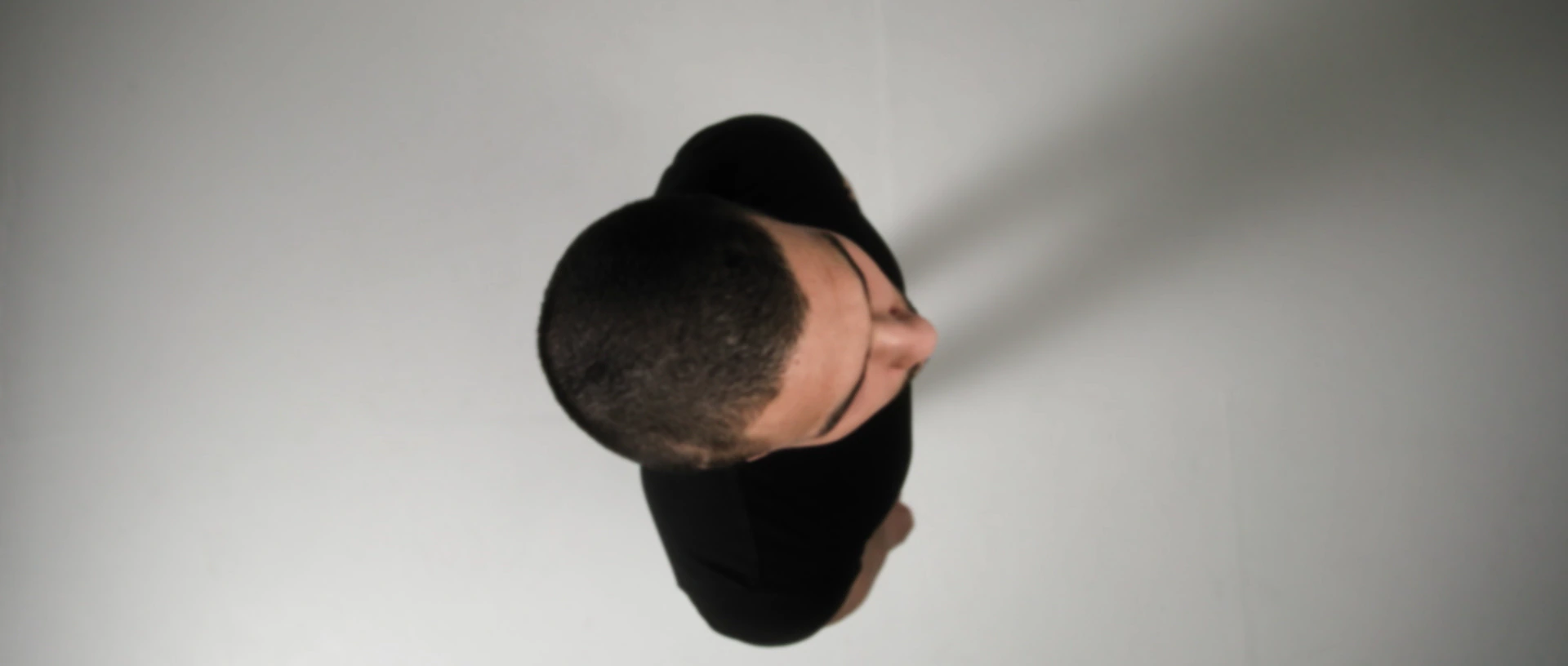 Un homme en t-shirt noir vu du dessus, sur un fond blanc où s'étire son ombre.