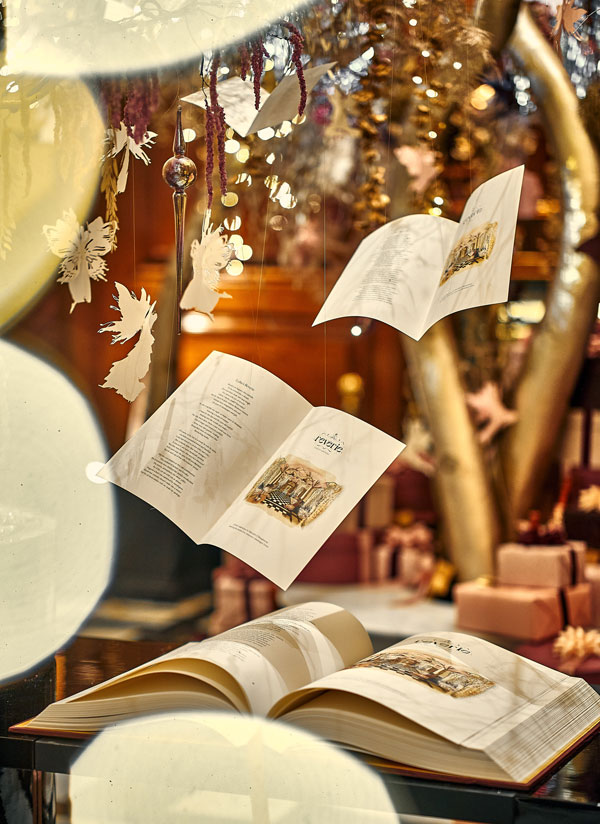 Open book with illustrated pages on a table, surrounded by hanging paper angels, ornaments, and blurred Christmas decorations in the background.