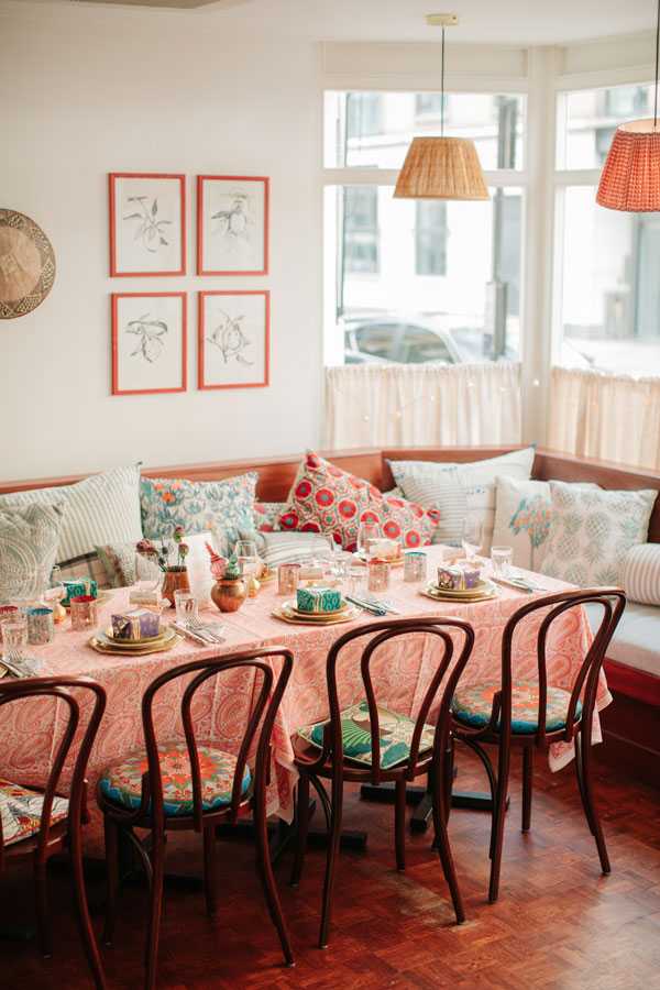 Cozy dining area with a table set for six, featuring colorful cushions, a patterned tablecloth, framed botanical prints, and hanging wicker lamps.