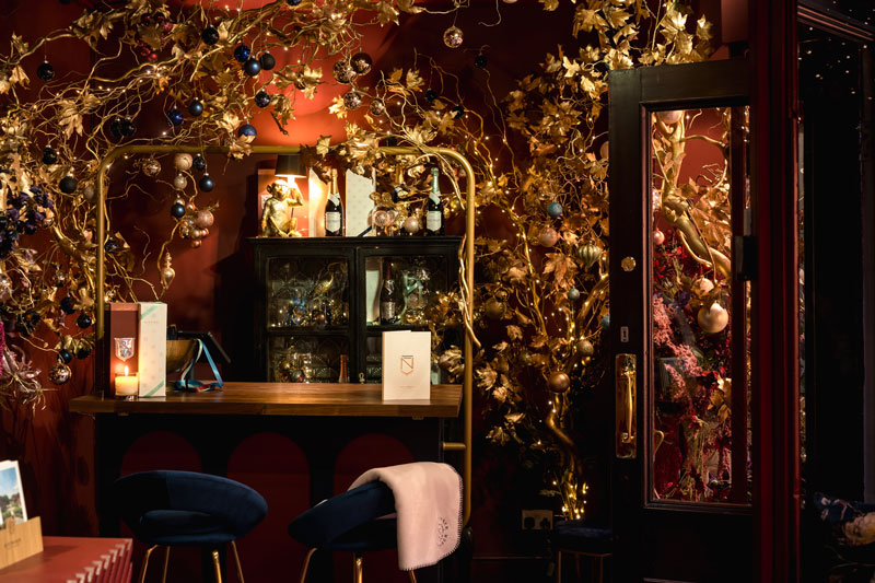 Cozy bar area with dark red walls, decorated with golden branches and hanging ornaments, featuring a wooden counter with two blue chairs and warm ambient lighting.