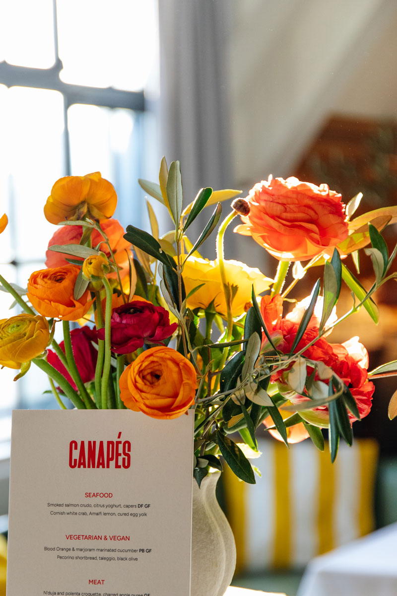 Vase of vibrant orange, yellow, and red ranunculus flowers next to a canapé menu card on a sunlit table.