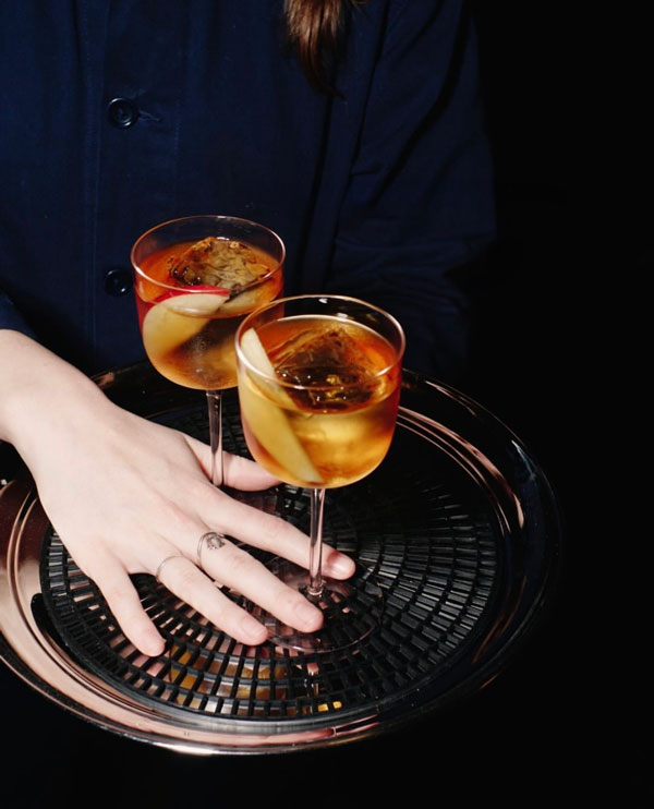 Two cocktail glasses with amber drinks and apple slices on a black tray held by a person wearing a dark outfit.