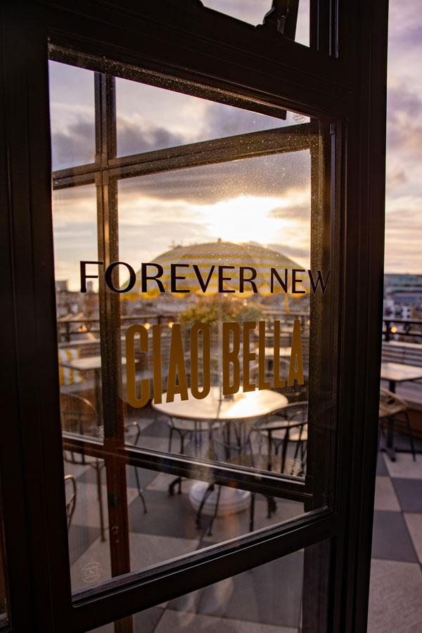 View through a glass door with 'FOREVER NEW CIAO BELLA' text, showing outdoor seating and sunset sky.