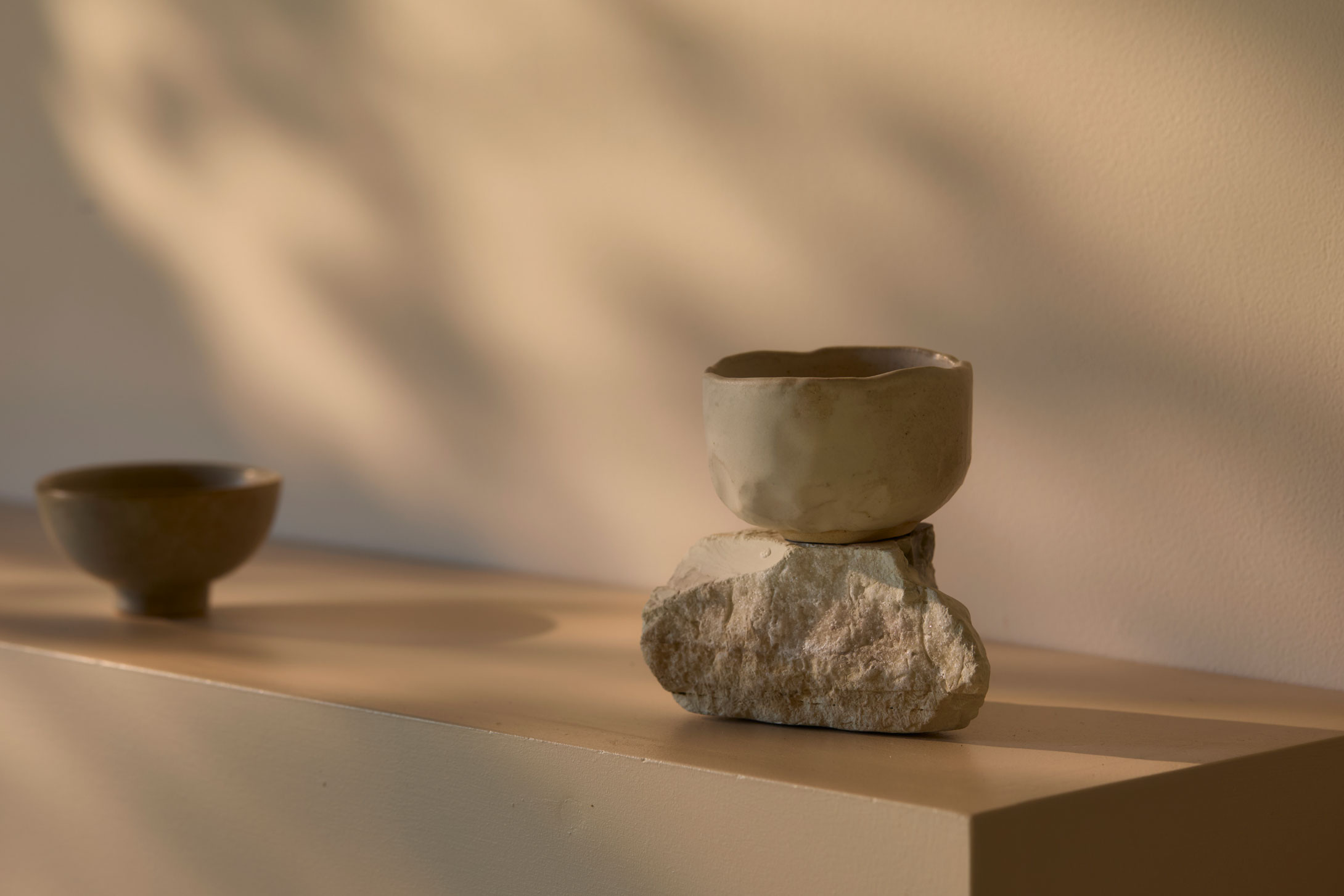 A handmade ceramic bowl placed on a rough stone on a beige shelf with soft shadowed light.