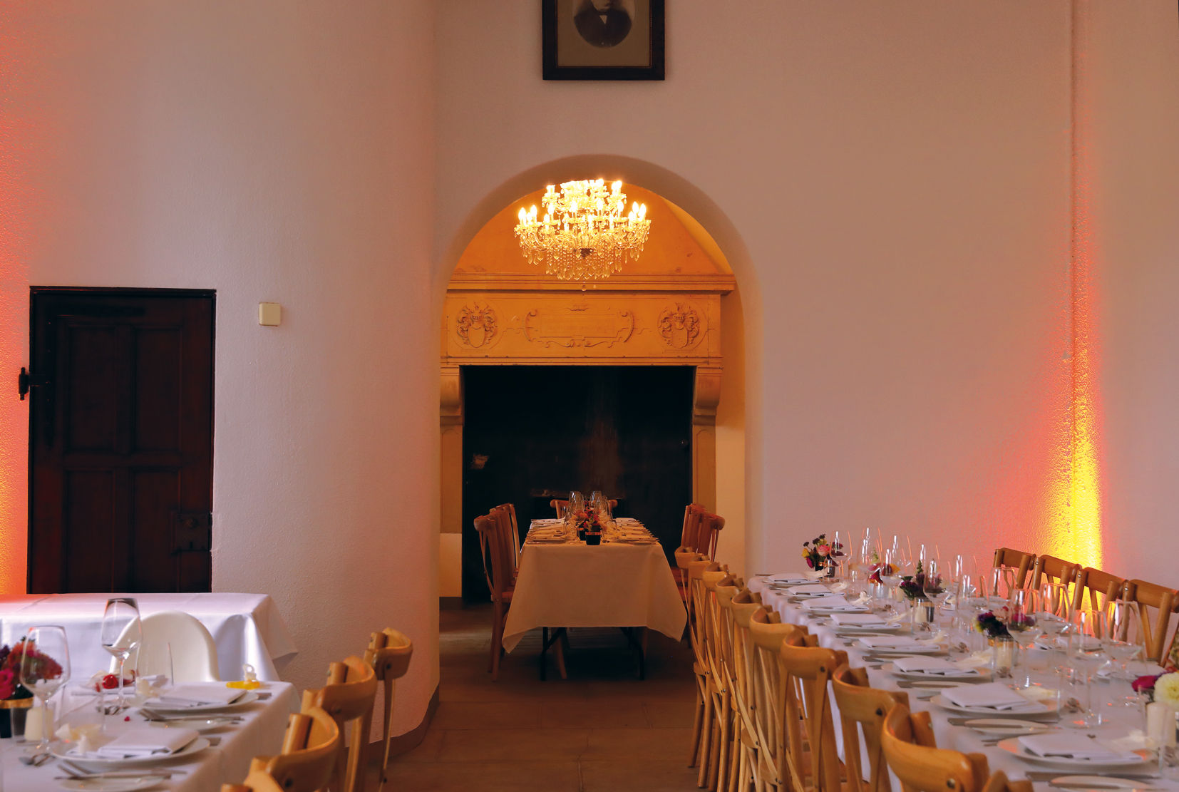 Festlich gedeckte Tische mit weißen Tischdecken und Holzstühlen in einem Raum mit Bogen, Kronleuchter und Kamin.