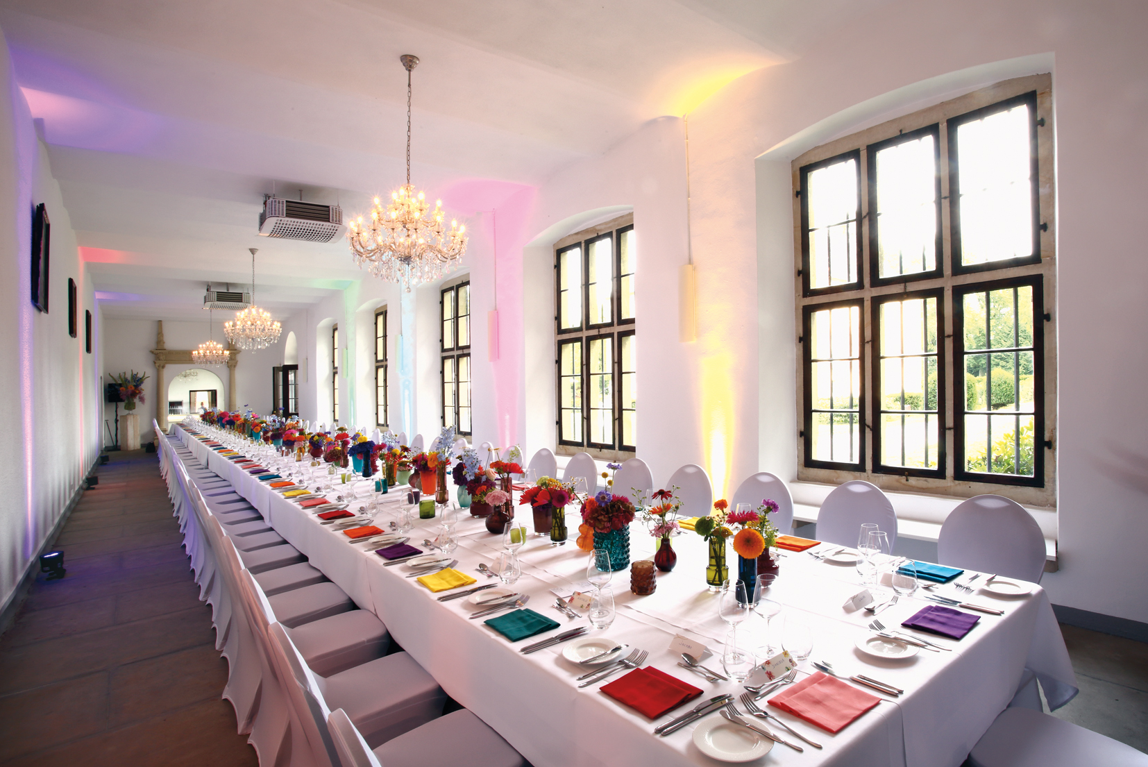 Langer festlich gedeckter Tisch mit weißen Stühlen, bunten Servietten und mehreren Blumenarrangements in einem hellen Raum mit großen Fenstern und Kronleuchtern.