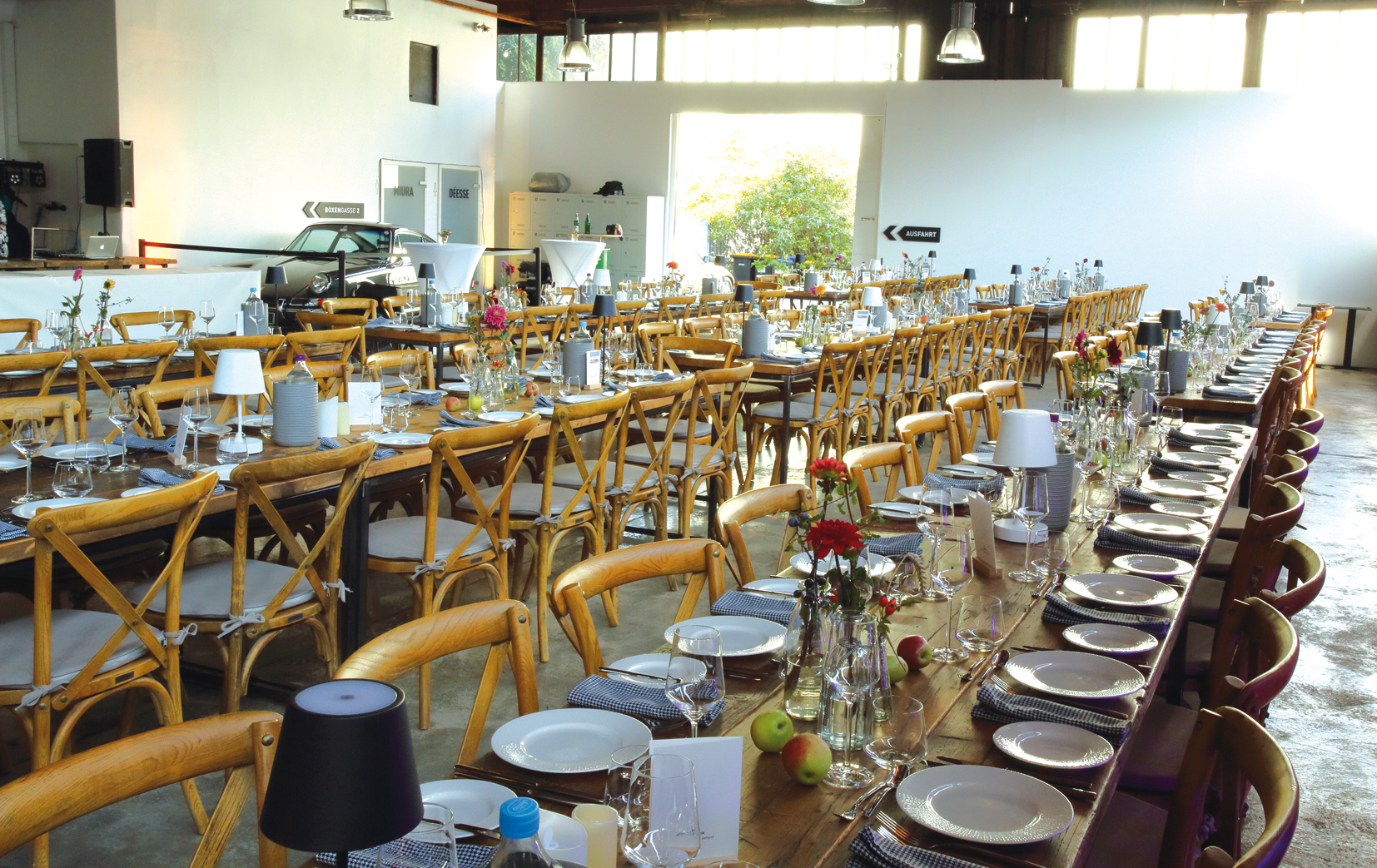 Großer Veranstaltungsraum mit langen Holztischen, gedeckt mit Tellern, Gläsern, Besteck, Blumen und Äpfeln auf den Tischen.