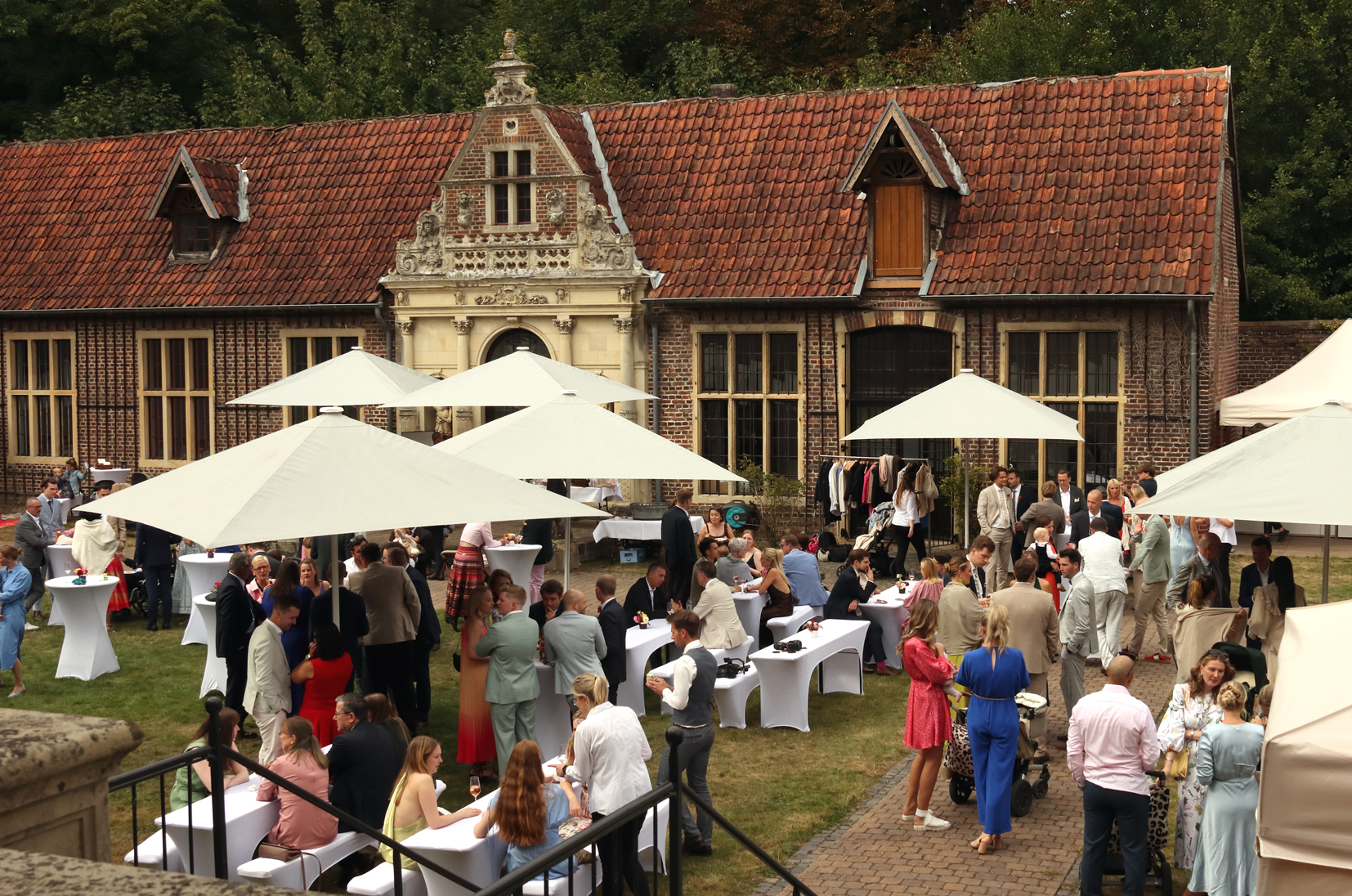 Draußenstehende Gruppe von Menschen, die sich unter weißen Sonnenschirmen bei einer Veranstaltung vor einem historischen Backsteingebäude mit rotem Ziegeldach unterhalten.
