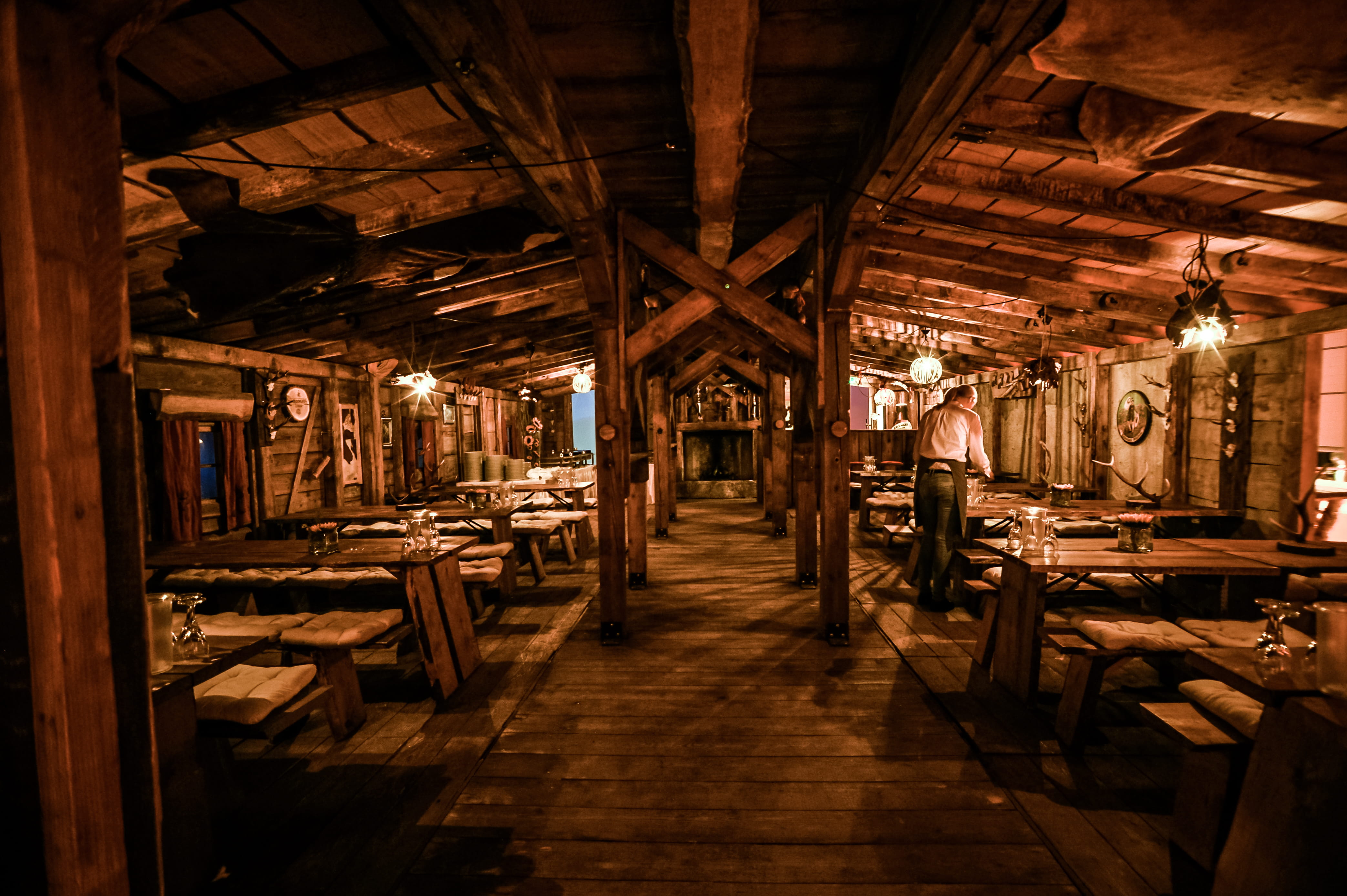 Rustikales Restaurant mit Holztischen und Sitzbänken, warm beleuchtet und einem Kellner, der einen Tisch vorbereitet.