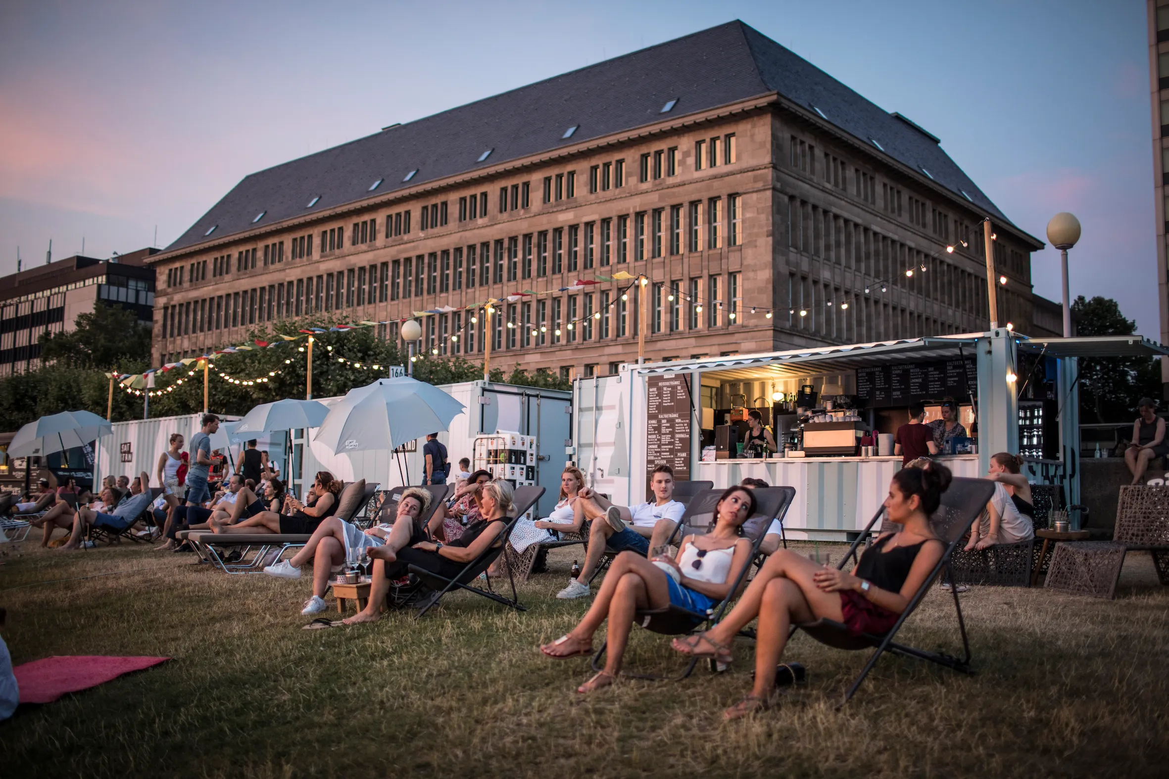 Menschen entspannen auf Liegestühlen auf einer Wiese vor einem großen Gebäude bei einem Freiluft-Café mit Lichterketten.
