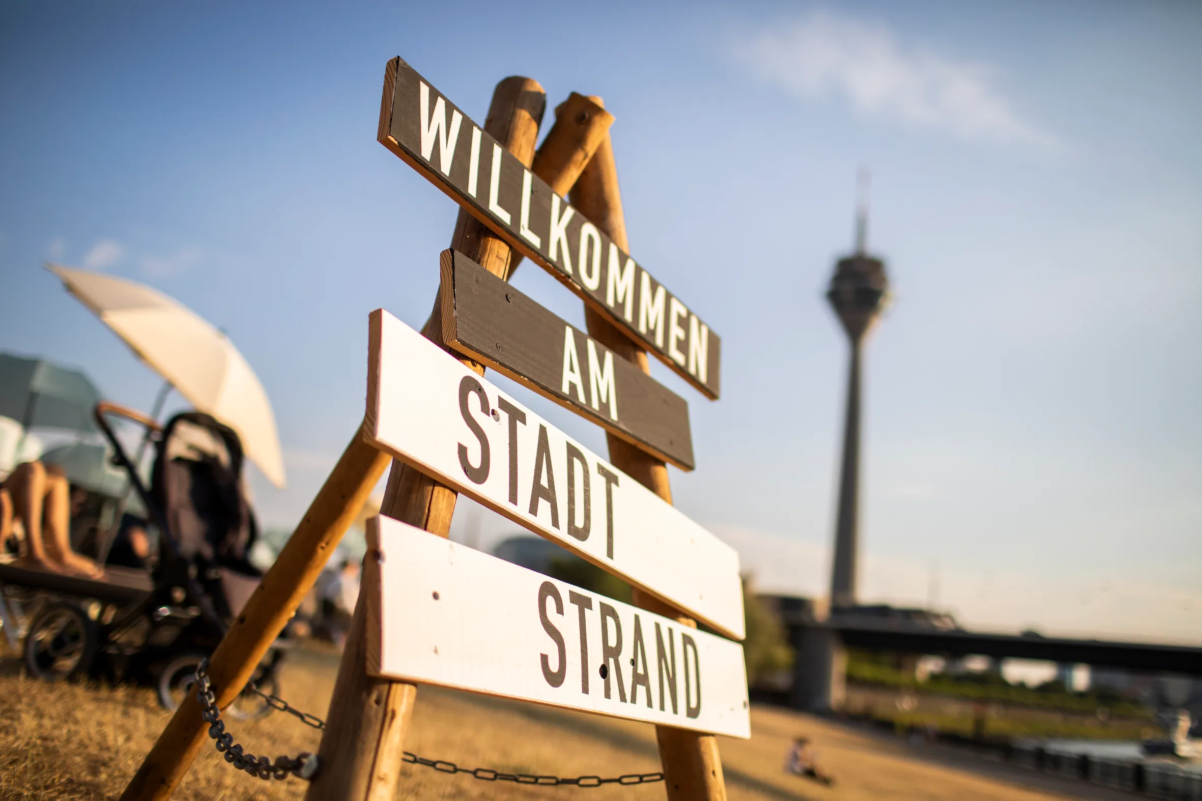 Holzschild mit der Aufschrift 'Willkommen am Stadtstrand' vor dem Rheinturm in Düsseldorf bei Sonnenschein.