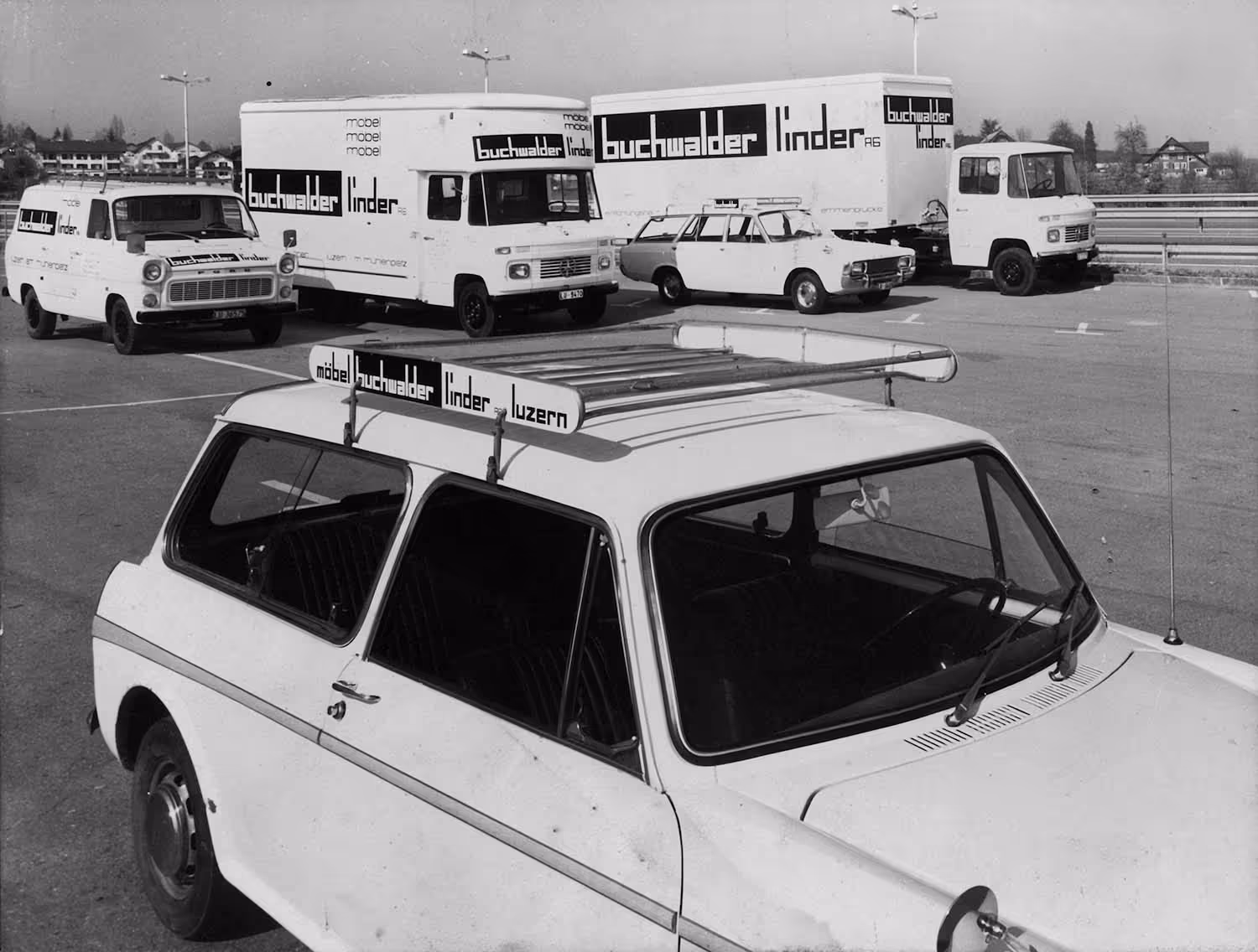 Mehrere weiße Fahrzeuge mit Aufschrift „buchwalder linder“ auf einem Parkplatz, darunter Lieferwagen, Kastenwagen und ein Auto mit Dachwerbung.