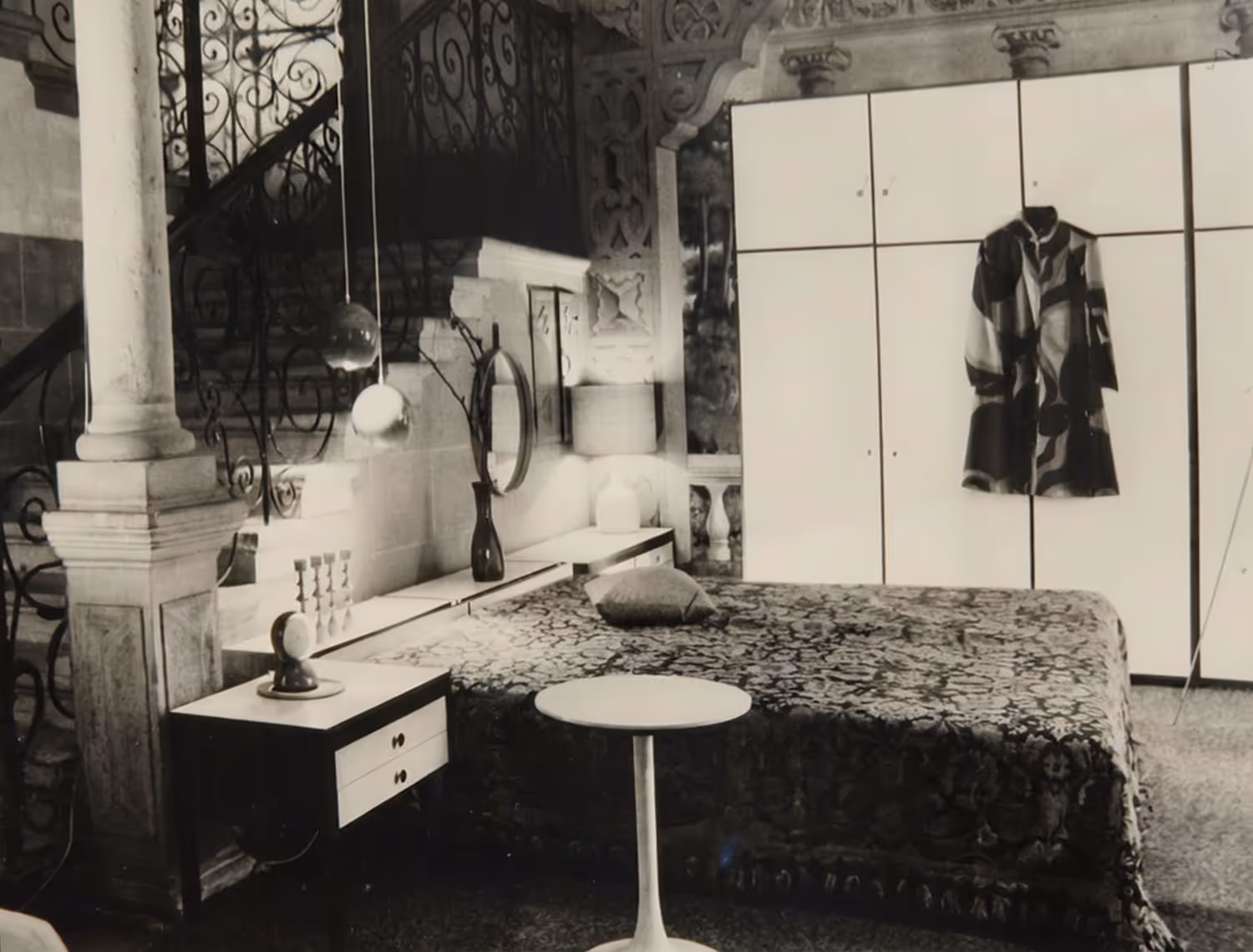 Schwarz-weisses Bild eines Schlafzimmers mit gemustertem Bettüberwurf, kleinem runden Tisch, Nachttisch mit Kerzenhalter und einem gemusterten Kleid, das an einem weissen Kleiderschrank hängt.
