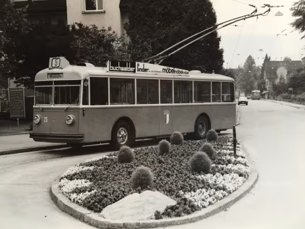 Schwarz-weiß-Foto eines Trolleybusses der Linie 5, der an einer mit Blumen und dekorativen Büschen bepflanzten Verkehrsinsel vorbeifährt.