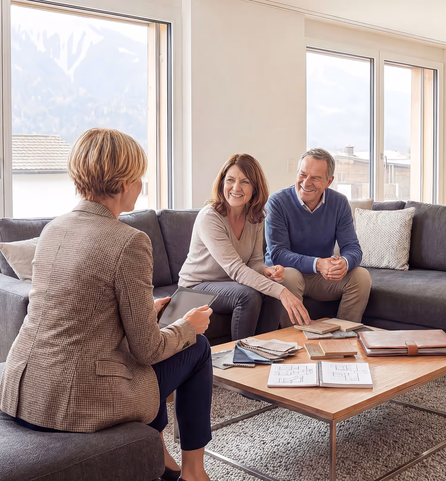 Drei Personen sitzen in einem modernen Wohnzimmer und besprechen Holzproben und Architektenpläne auf einem Couchtisch.