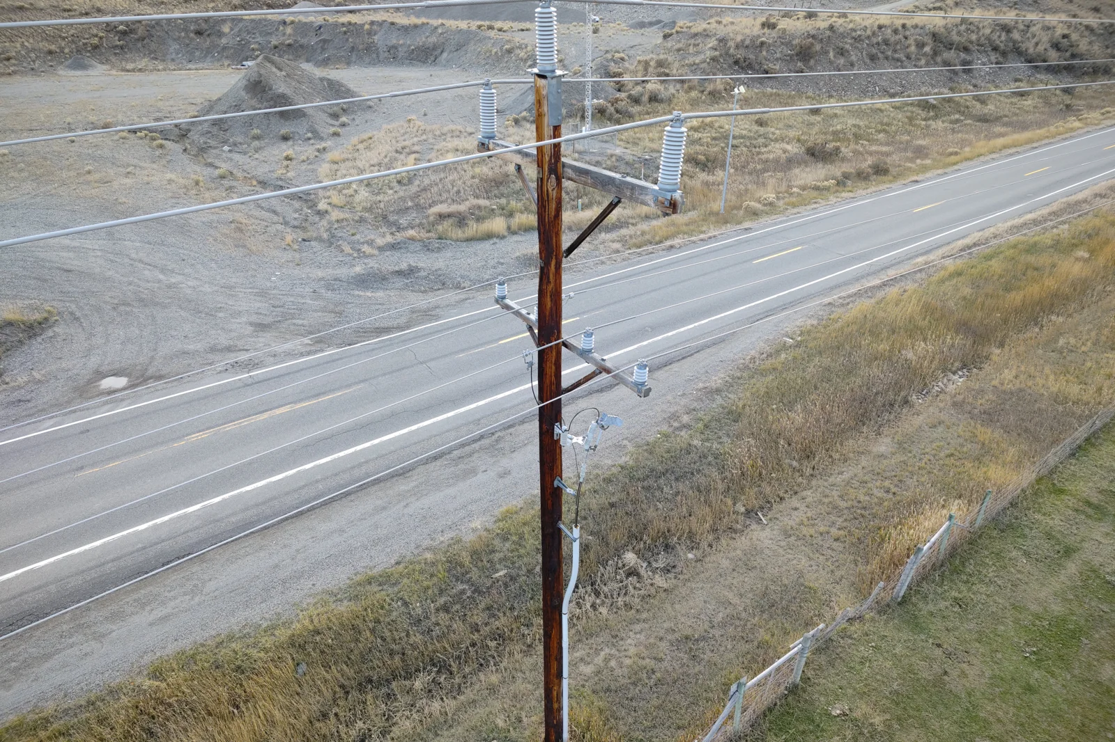 Drone-altitude view of a wooden distribution pole beside a highway, showing crossarm hardware a ground patrol sees only as a silhouette