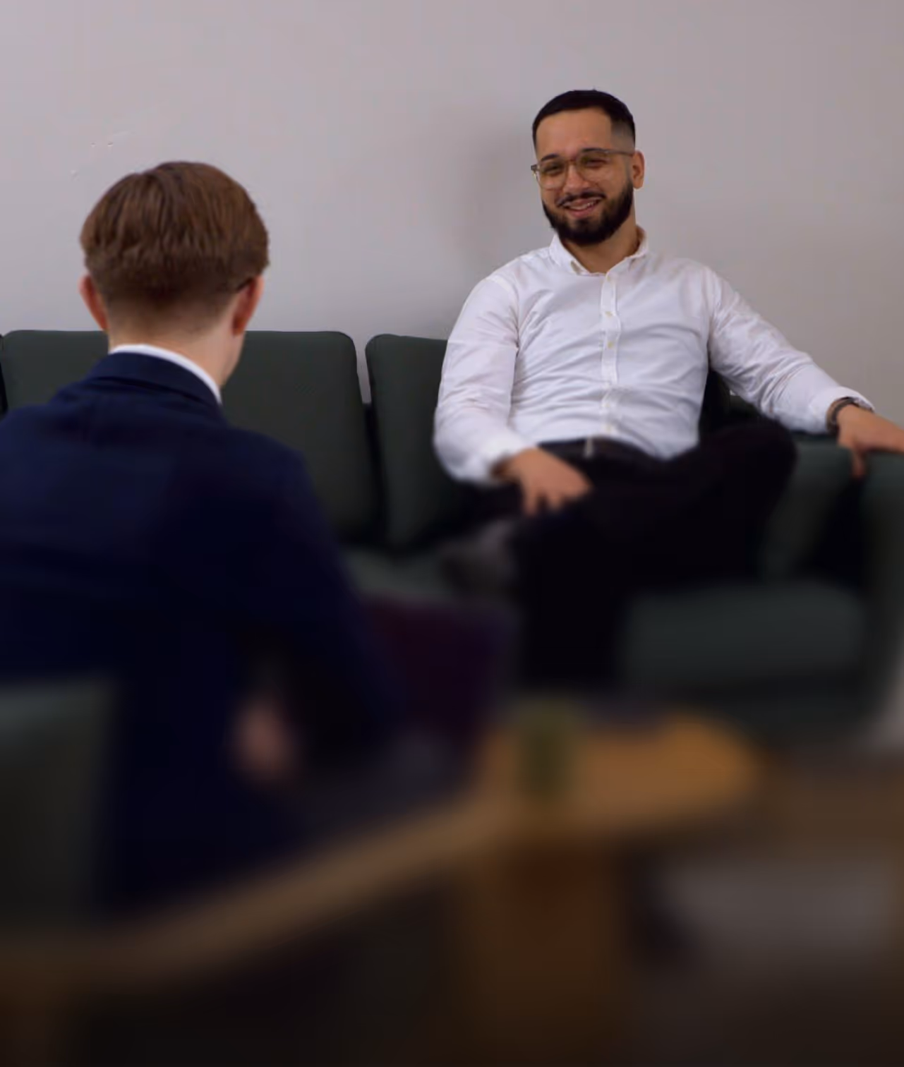 Man with glasses and white shirt sitting on a green couch, smiling during a conversation with another man in a navy suit seen from behind.