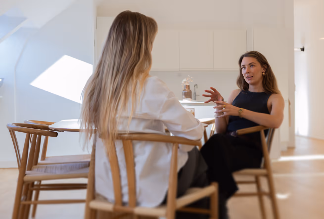 Two women sitting on wooden chairs in a bright room, one talking and gesturing with her hands.
