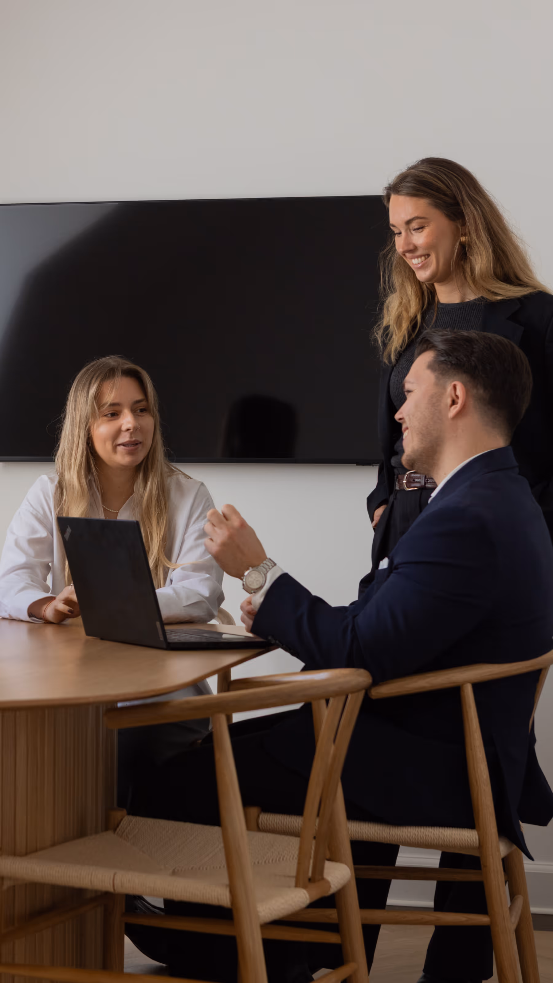 Four colleagues having a discussion around a wooden table with a laptop and a small vase with flowers, in a modern office setting.