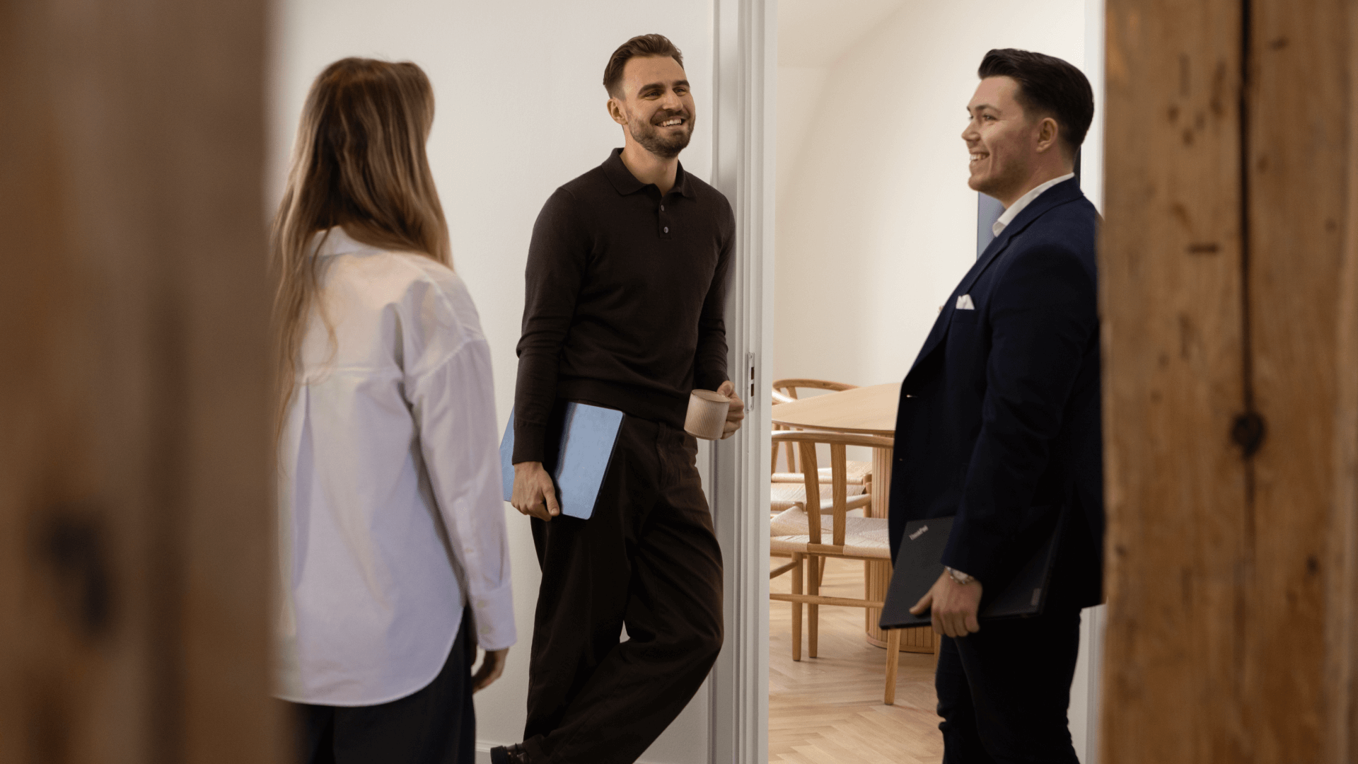 Three colleagues smiling and chatting in an office doorway, one holding a laptop and a coffee cup.