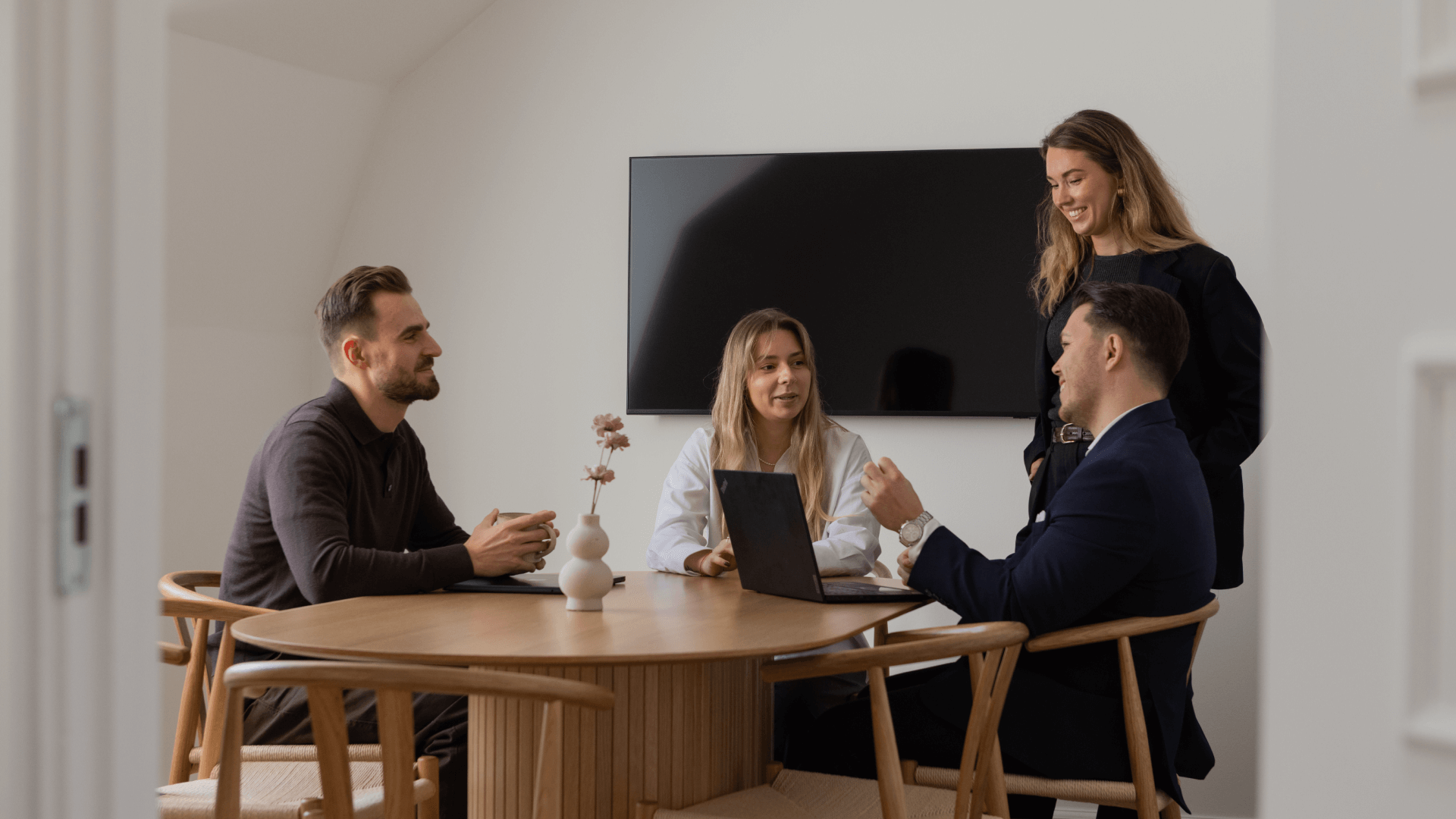 Four colleagues having a discussion around a wooden table with a laptop and a small vase with flowers, in a modern office setting.