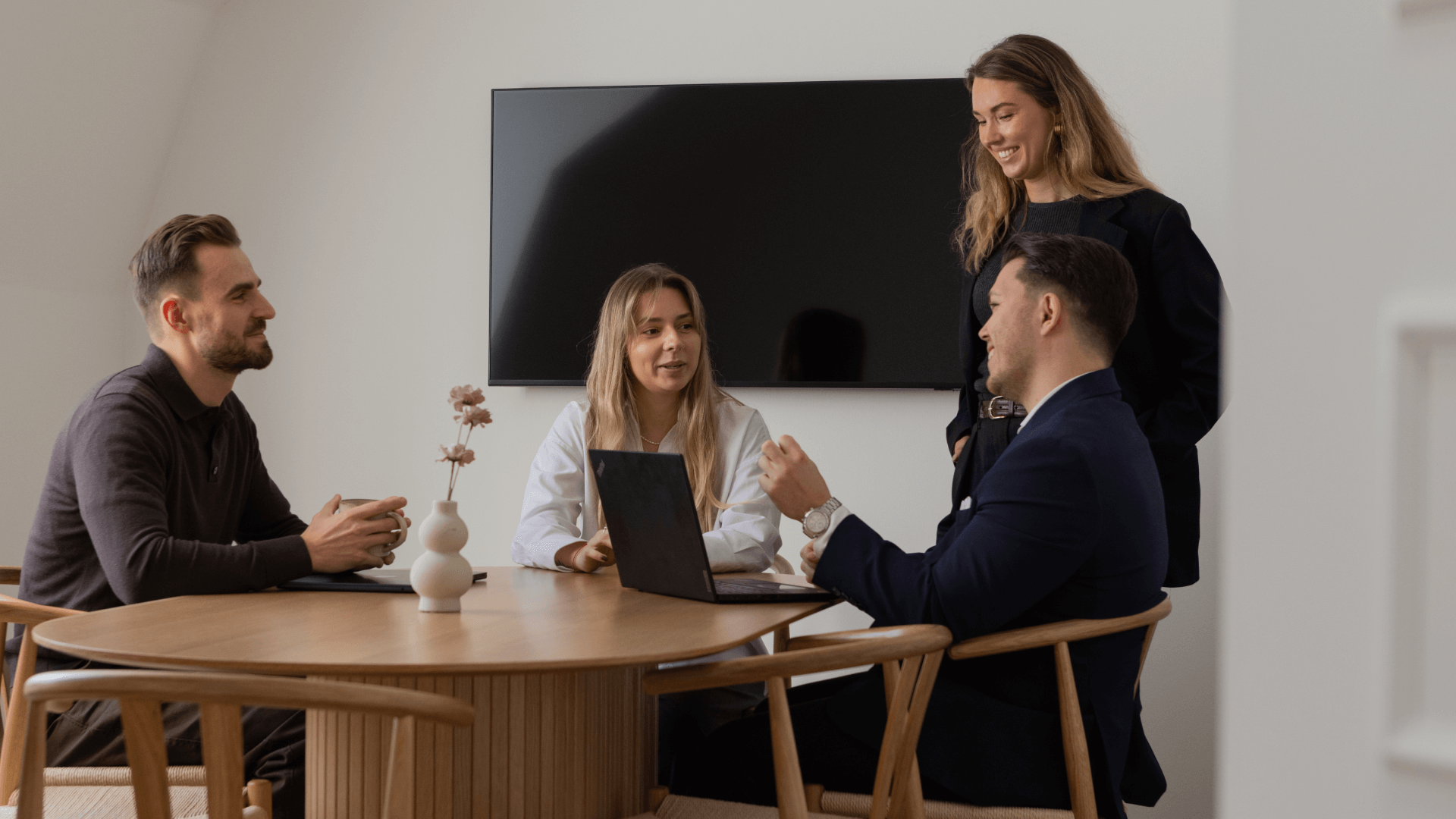 Four colleagues engaged in a meeting around a wooden table with a laptop and a TV screen in a modern office.