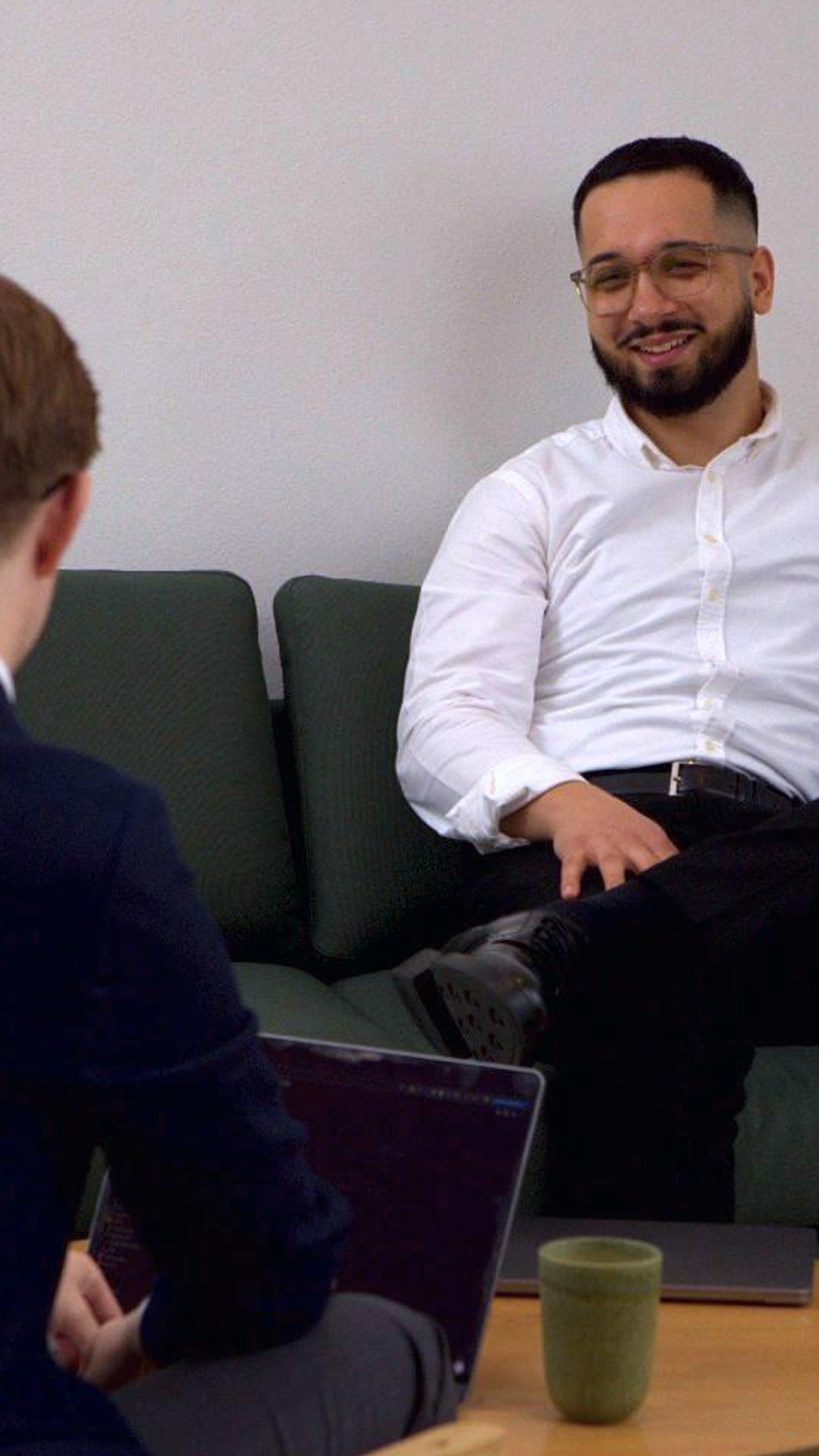 Two men having a conversation across a table, one smiling and wearing a white shirt, the other using a laptop.