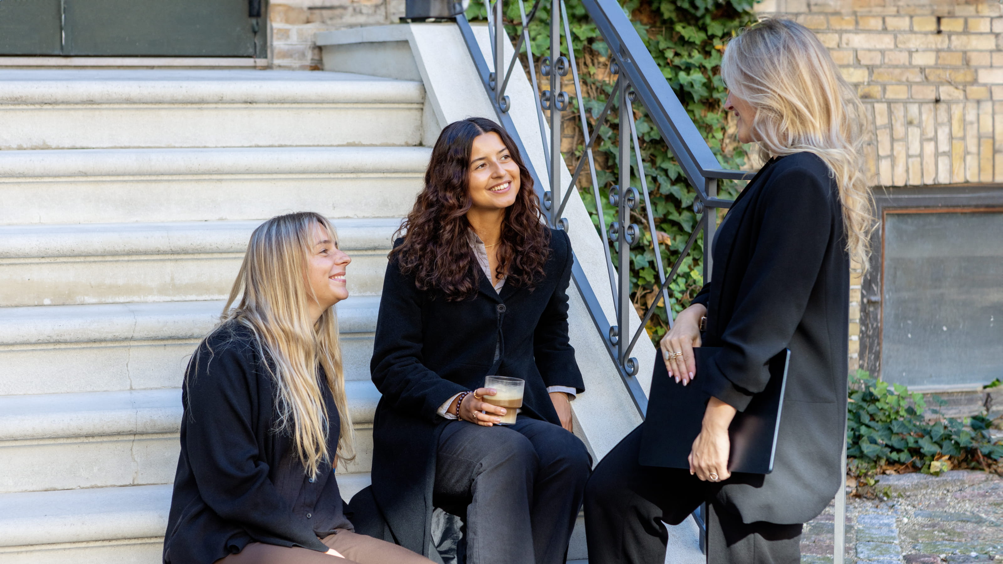 Three women in black coats chatting and smiling on outdoor stone steps, one holding a glass of coffee.