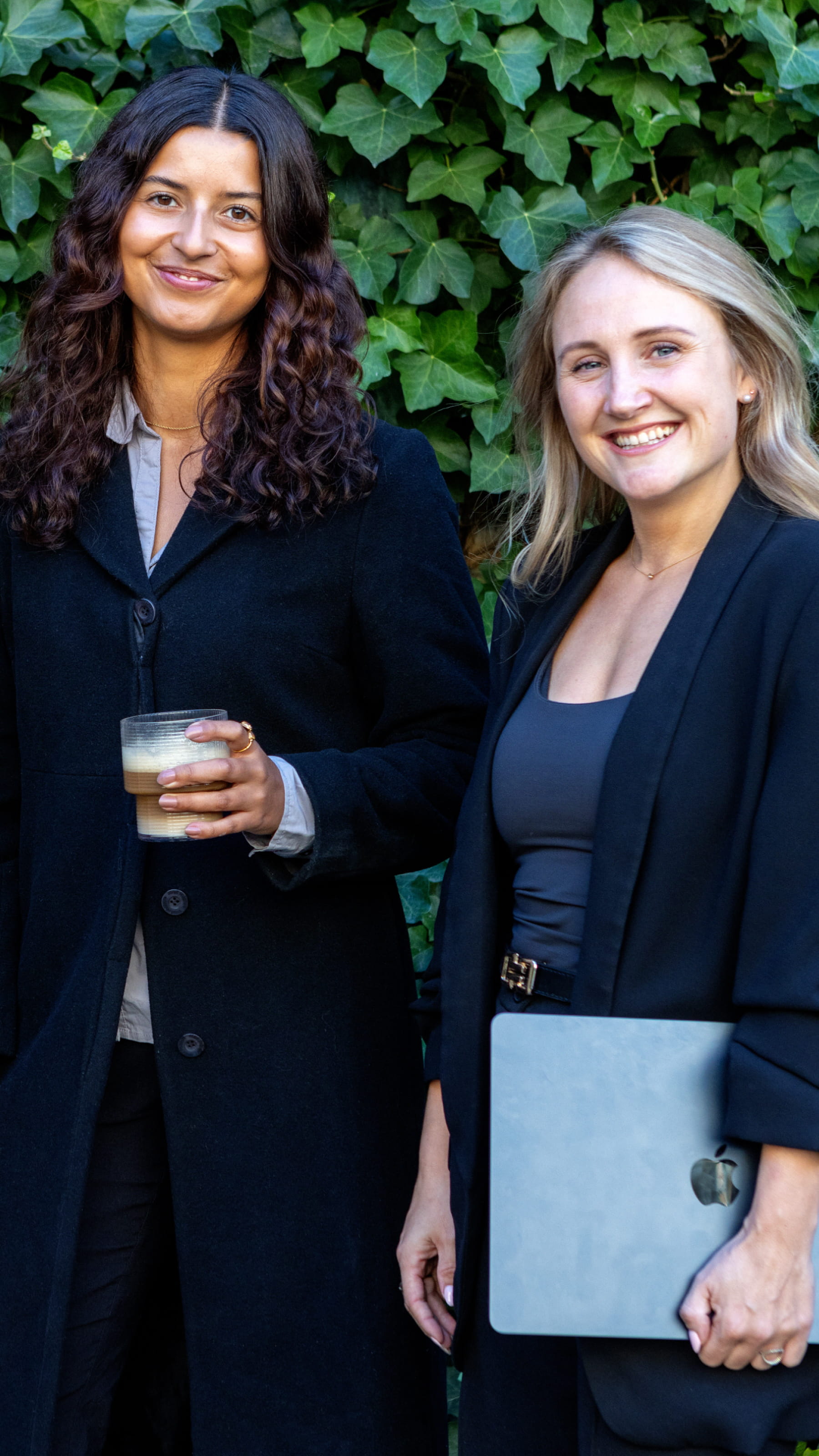 Two smiling women standing in front of green ivy, one holding a drink and the other holding a silver Apple laptop.
