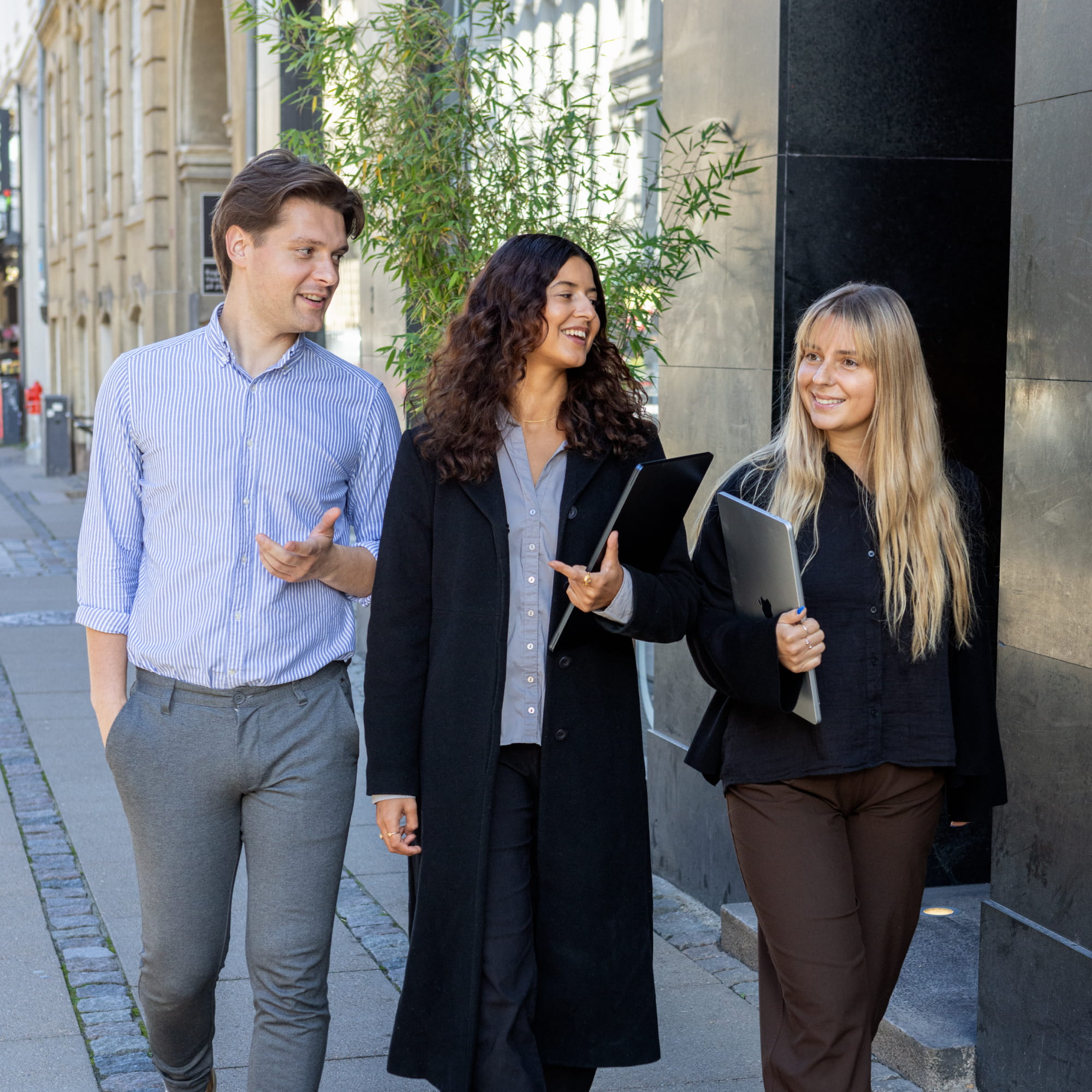 Three young professionals walking and talking on a city sidewalk, two women holding laptops.