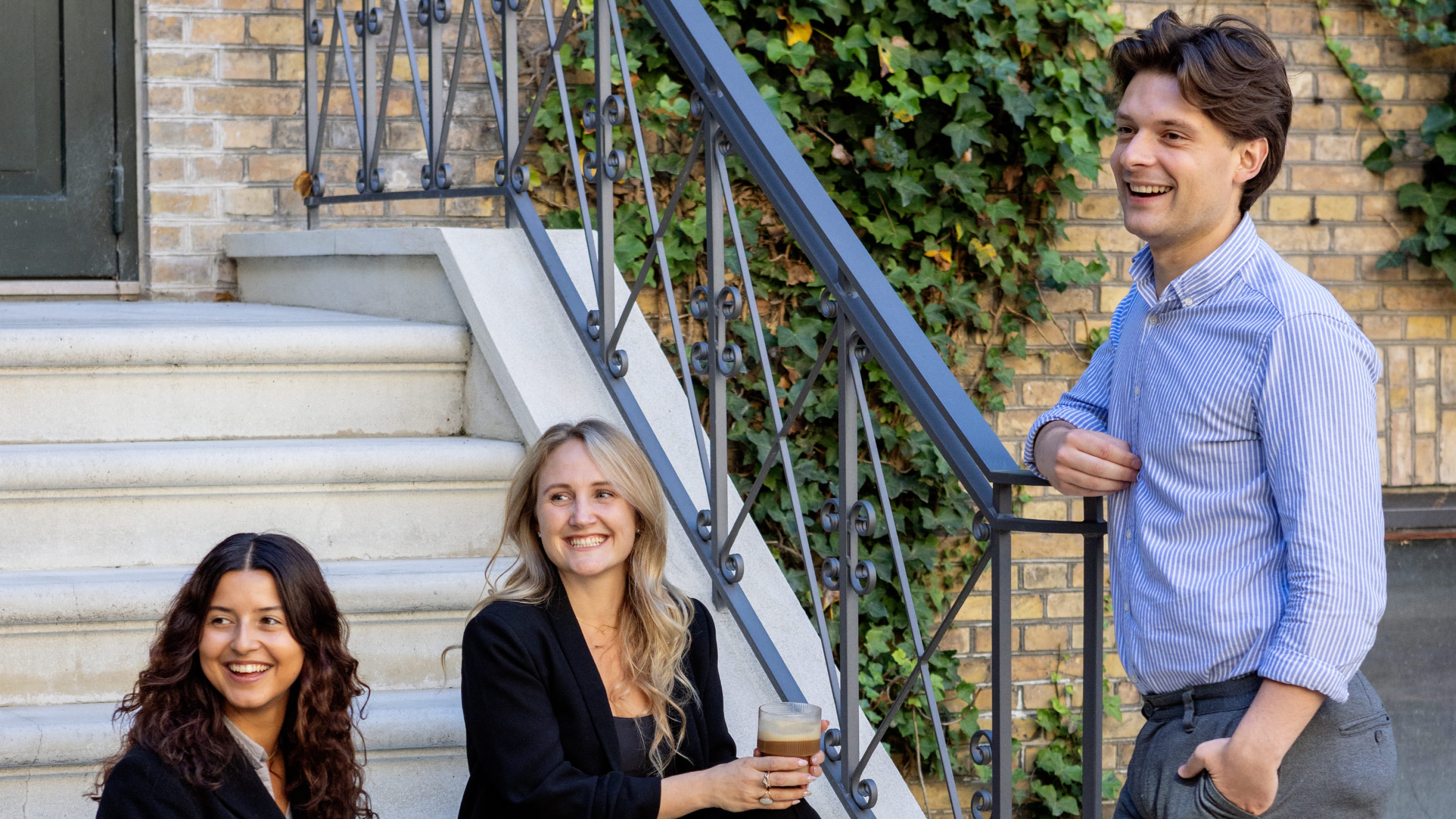 Two women sitting on outdoor stairs and a man standing nearby, all smiling and engaged in conversation.