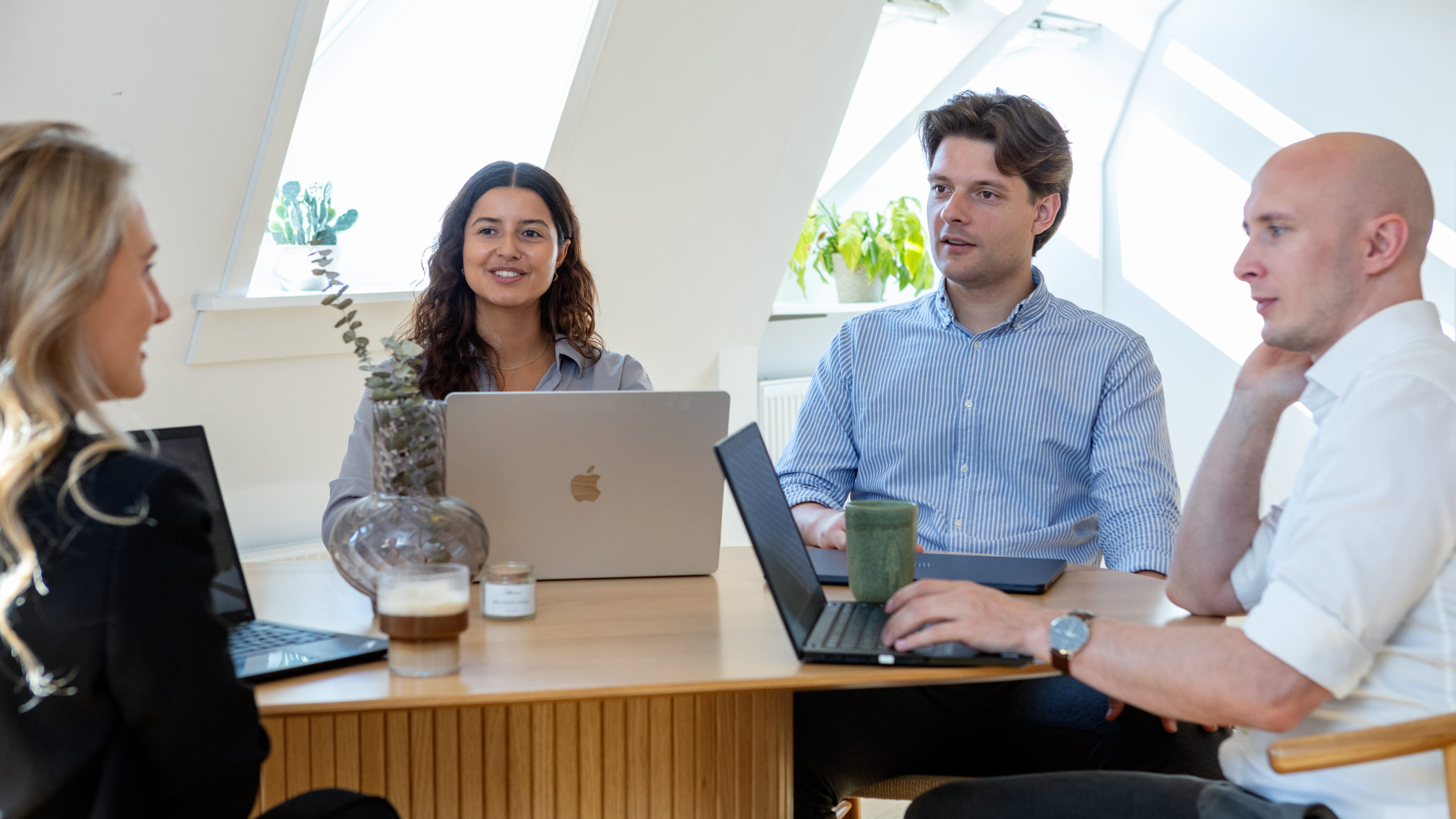 Four people having a discussion around a wooden table with laptops and drinks in a bright office space with skylights.