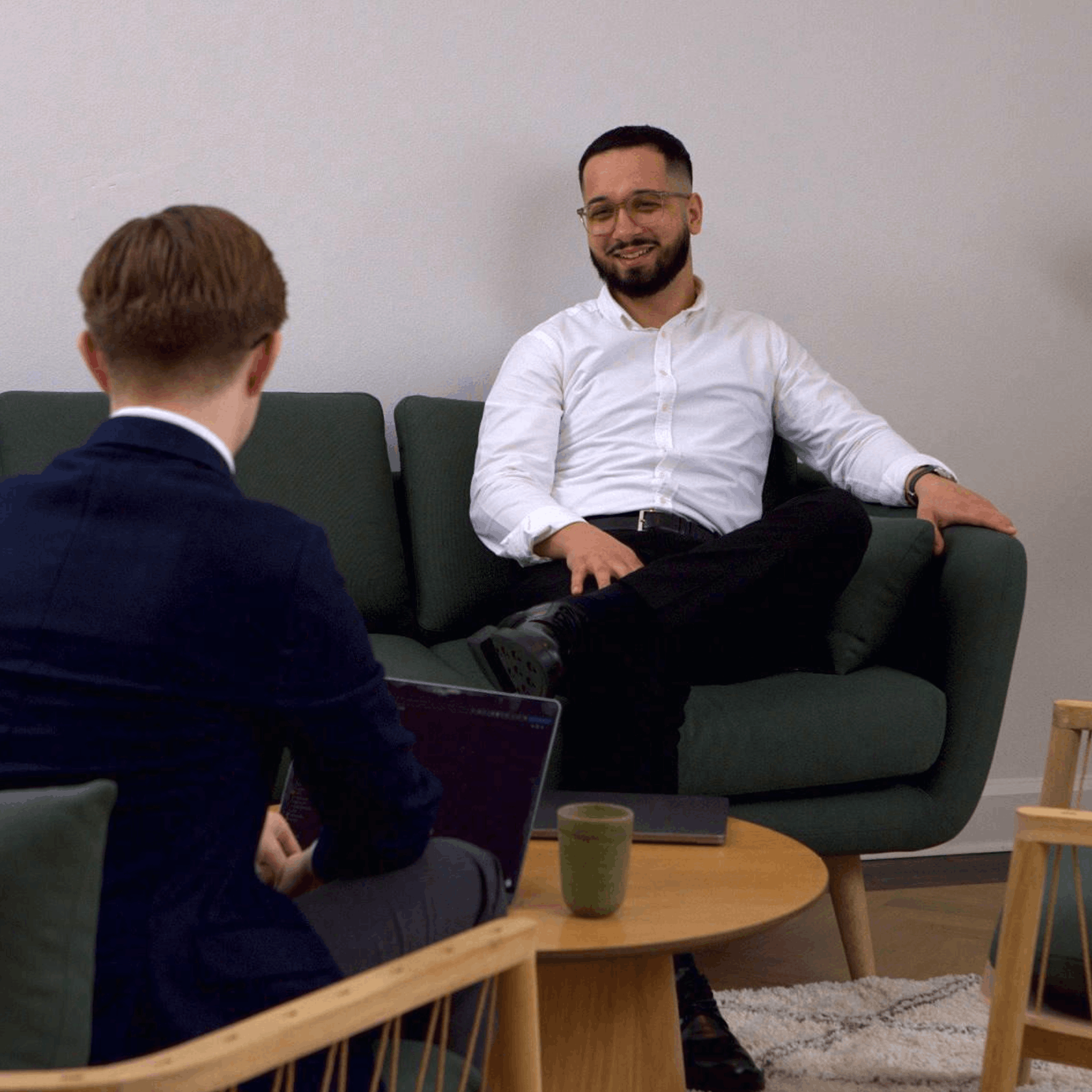 Man with glasses and white shirt sitting on a green couch, smiling during a conversation with another man in a navy suit seen from behind.