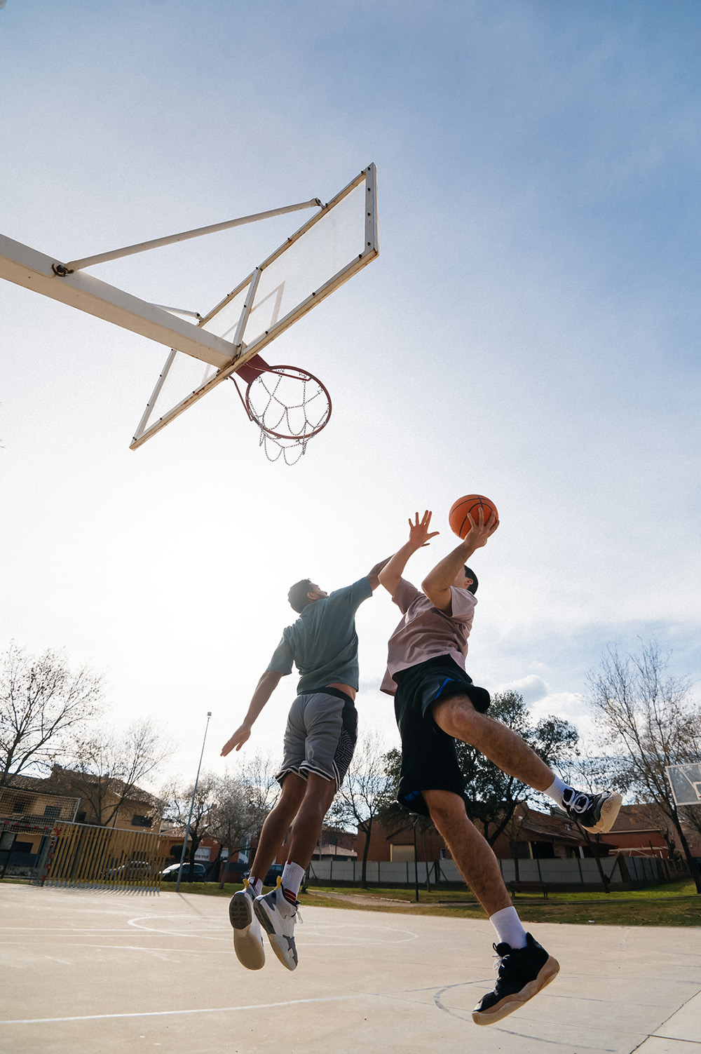 Two men jumping on an outdoor basketball court, one holding a basketball near the hoop, the other attempting to block.