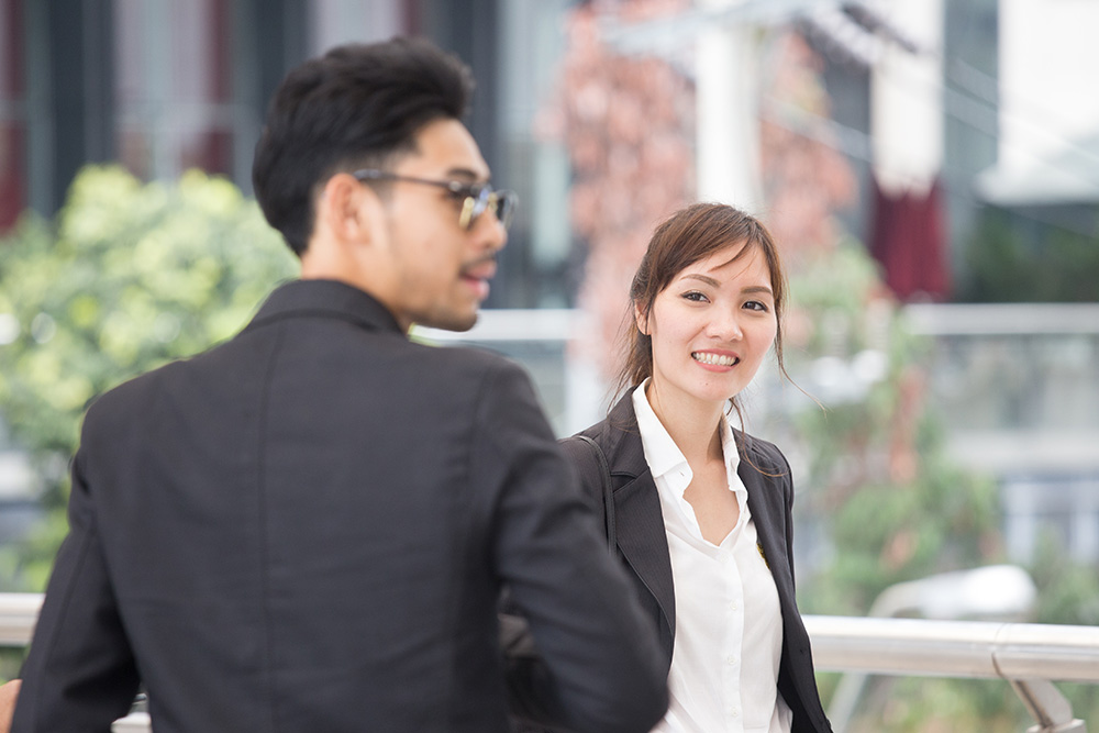 Two business professionals outdoors, a woman smiling and facing the camera while a man in sunglasses stands in the foreground.