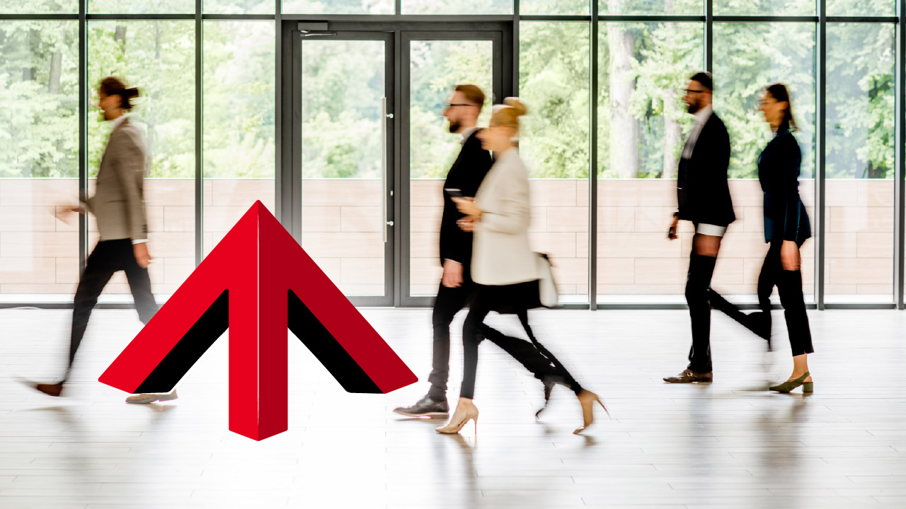 Two men and a woman in business attire engaged in a serious discussion in a modern office lounge area with others working at a table in the background.