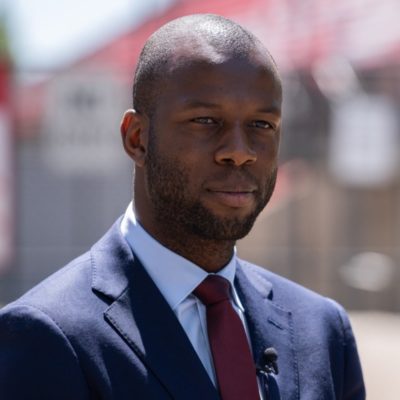 Photo of Bruce Djite in a blue suit and red tie standing outdoors with blurred buildings in the background. Client Testimonial.