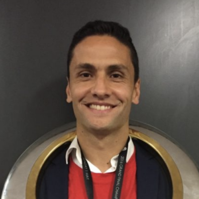 Photo of Marcelo Carrusca, wearing a red shirt, white collar, and black jacket, standing in front of a dark background with a circular metallic frame. Client Testimonial.
