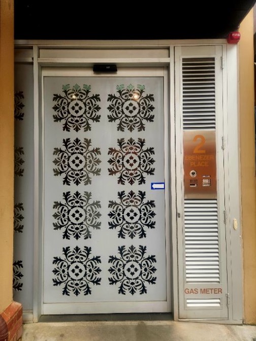 Photo of the Archer Head Office front door, a gray door with eight symmetrical black decorative patterns arranged in a grid, next to a vented panel with a sign reading '2 Ebenezer Place'.