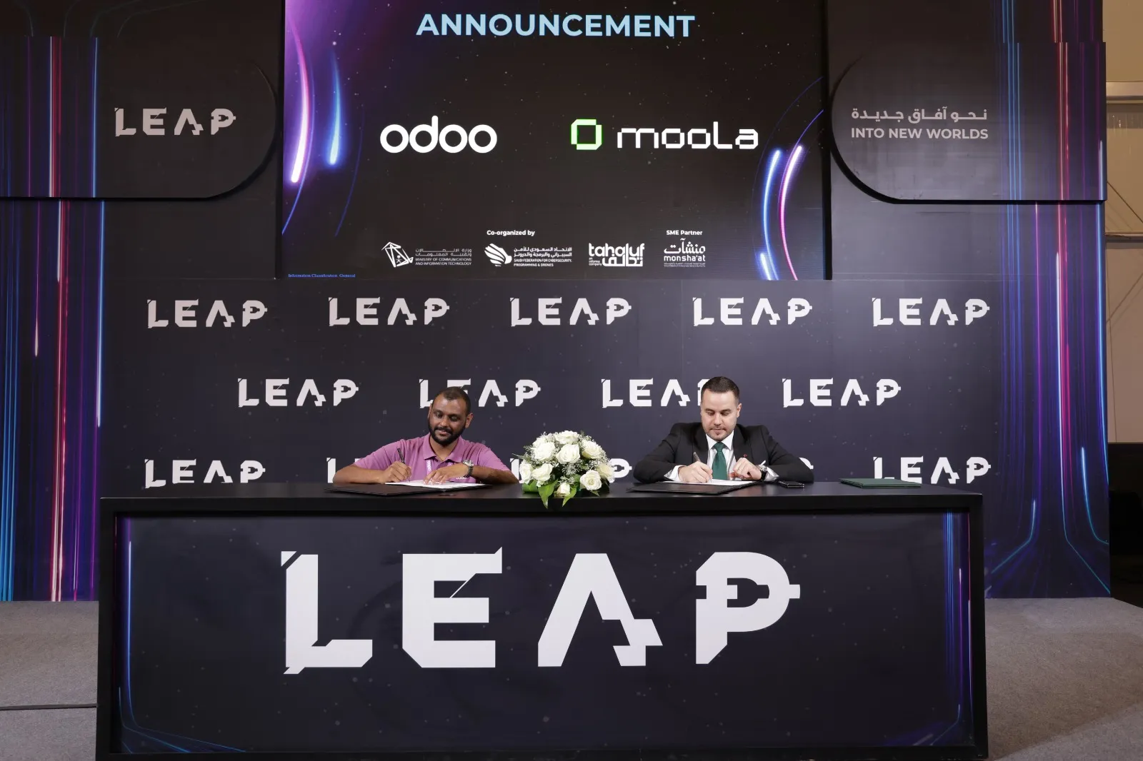 Two men seated at a table with LEAP branding, signing documents beneath a screen showing logos of Odoo and Moola.