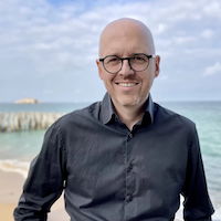 Homme chauve avec lunettes souriant, portant une chemise noire, devant une plage avec mer et ciel nuageux.