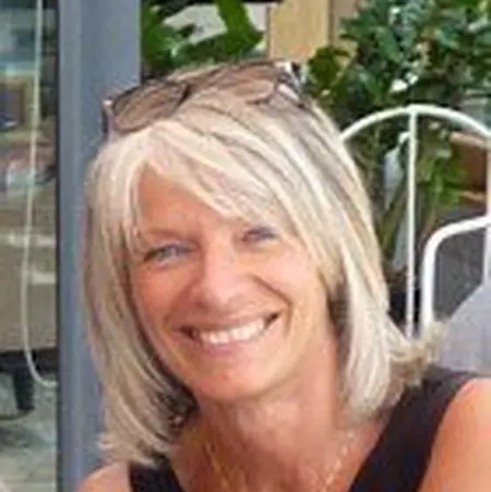 Femme souriante aux cheveux blonds portant des lunettes posées sur la tête, en extérieur près de plantes.